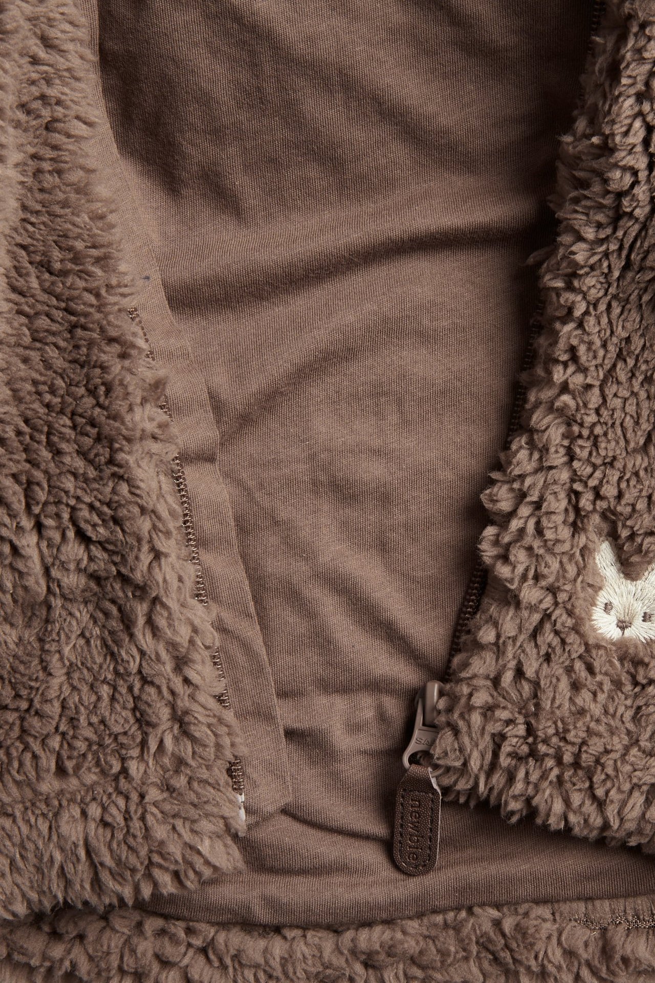 Nahaufnahme einer braunen flauschigen Babyjacke mit Stickerei und Reißverschluss über braunem Oberteil.