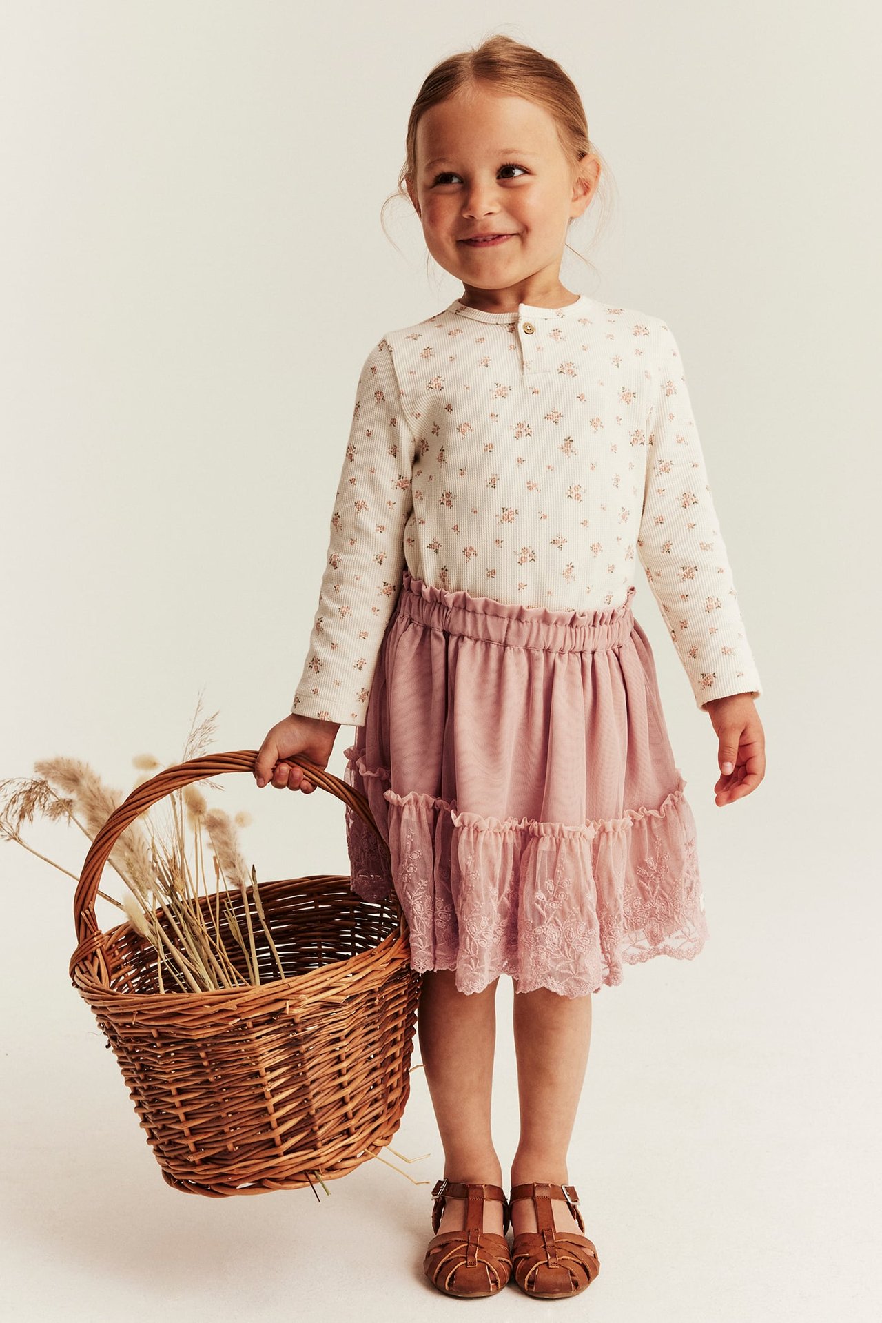 Vorderansicht: Kind in cremefarbenem Blümchen-Oberteil, pinkem Stufenrock, braunen Sandalen, hält Korb.
