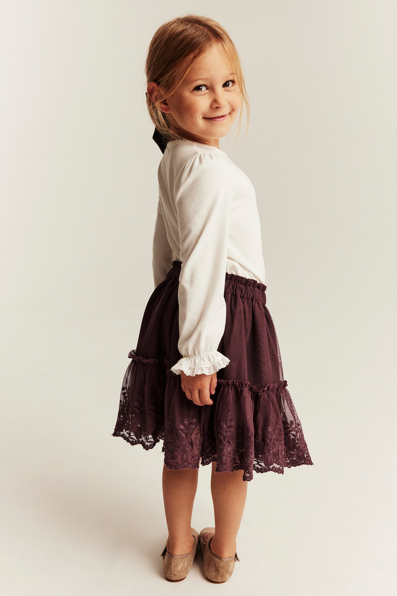 Mädchen in dunkelrotem Spitzenrock, weißem Langarmshirt mit Rüschen, braunen Schuhen, Seitenansicht.