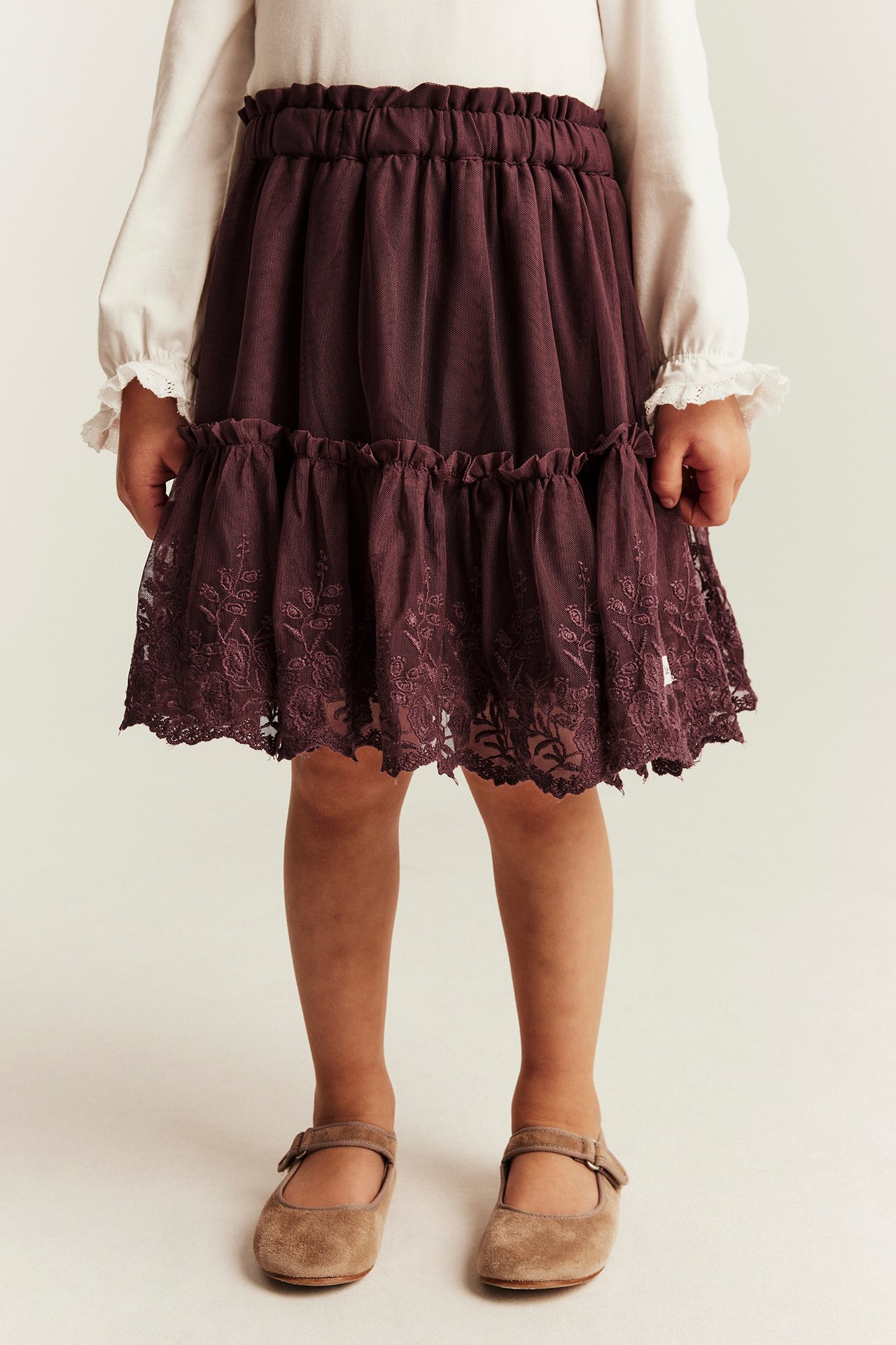 Front view of a kid wearing a dark purple tiered skirt with lace, a white top, and brown Mary Jane shoes.