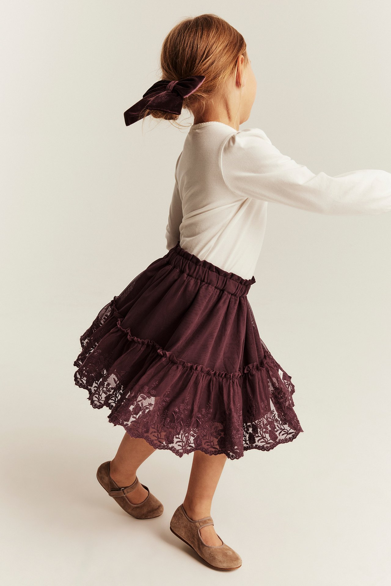 Back view: kid in a plum tiered lace skirt, white long-sleeve top, plum velvet bow, and brown Mary Jane flats.