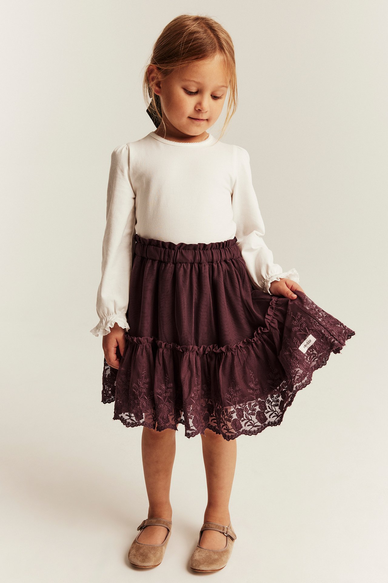 Vorderansicht eines Kindes in lila Stufenrock mit Spitze, weißem Langarmshirt und beigen Schuhen.