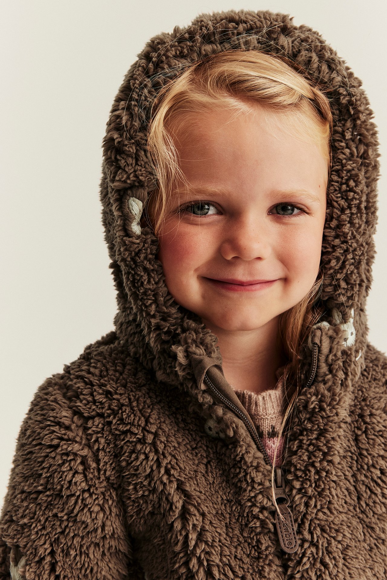 Nahaufnahme: Kind trägt braune Teddyfleece-Kapuzenjacke mit Stickerei über rosa Strickpullover.