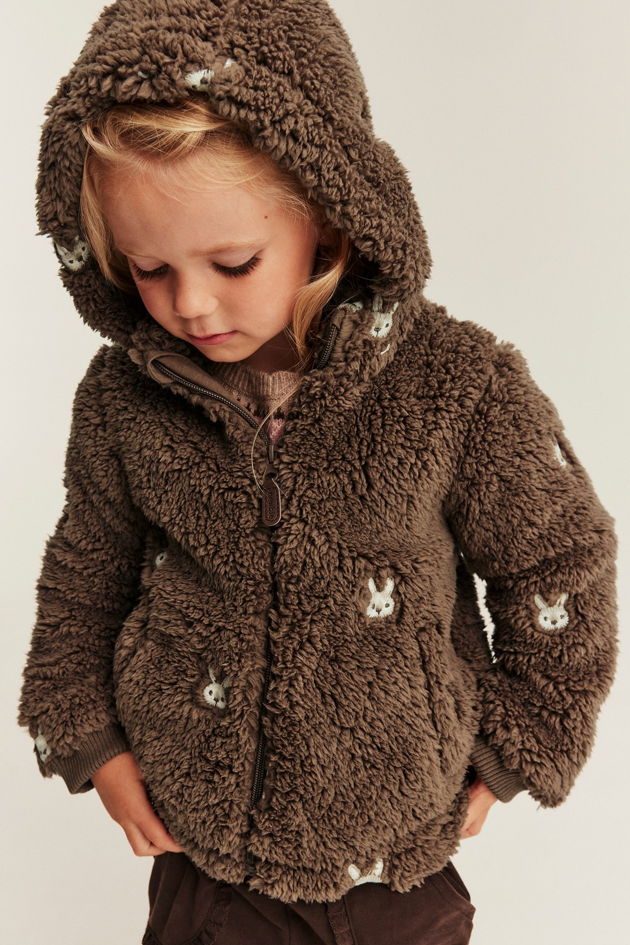 Front view: Kid wears a brown fluffy hooded jacket with bunny embroidery over a light brown sweater.