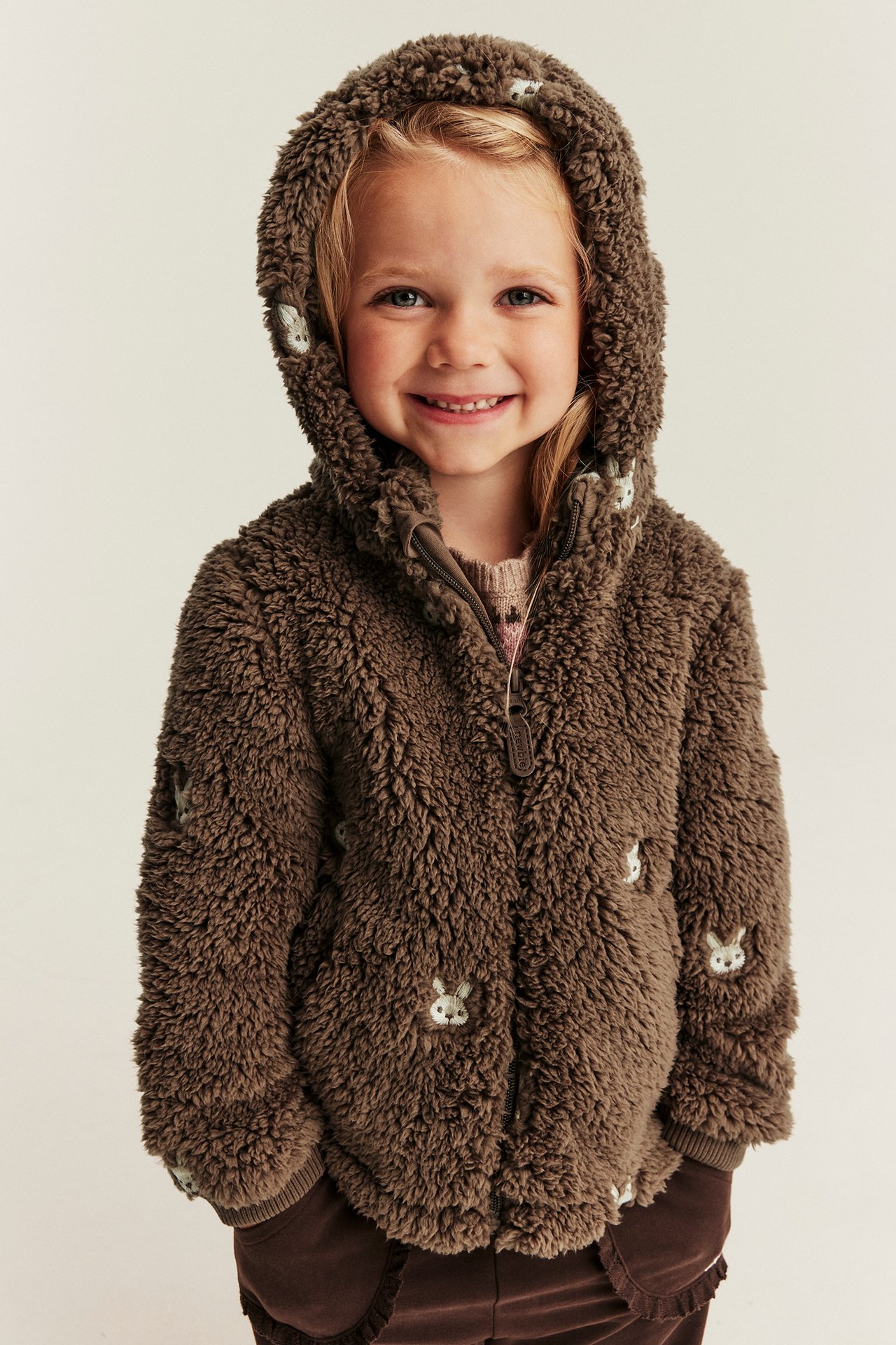 Vorderansicht von Kind in brauner flauschiger Kapuzenjacke mit Hasenmuster, rosa Oberteil und braunen Hosen.