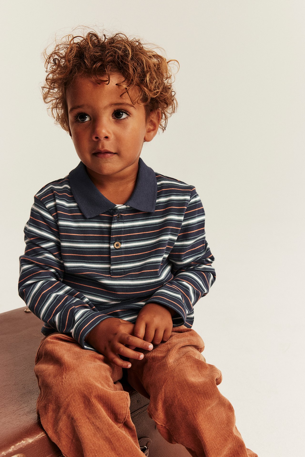 Vorderansicht: Kind in dunkelblau-gestreiftem langärmeligem Poloshirt und brauner Cordhose, sitzend.