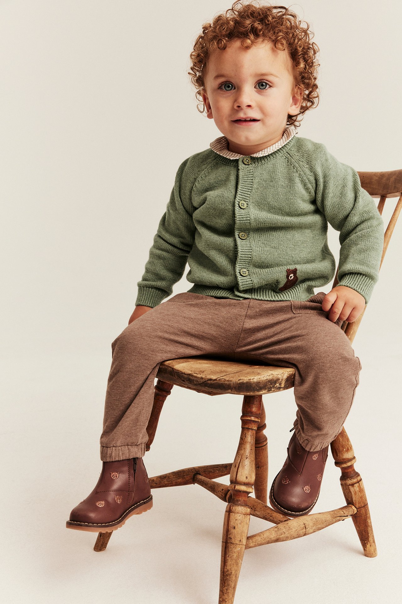 Vorderansicht eines Babys in grünem Cardigan mit Bärenapplikation, brauner Hose und braunen bestickten Stiefeln.