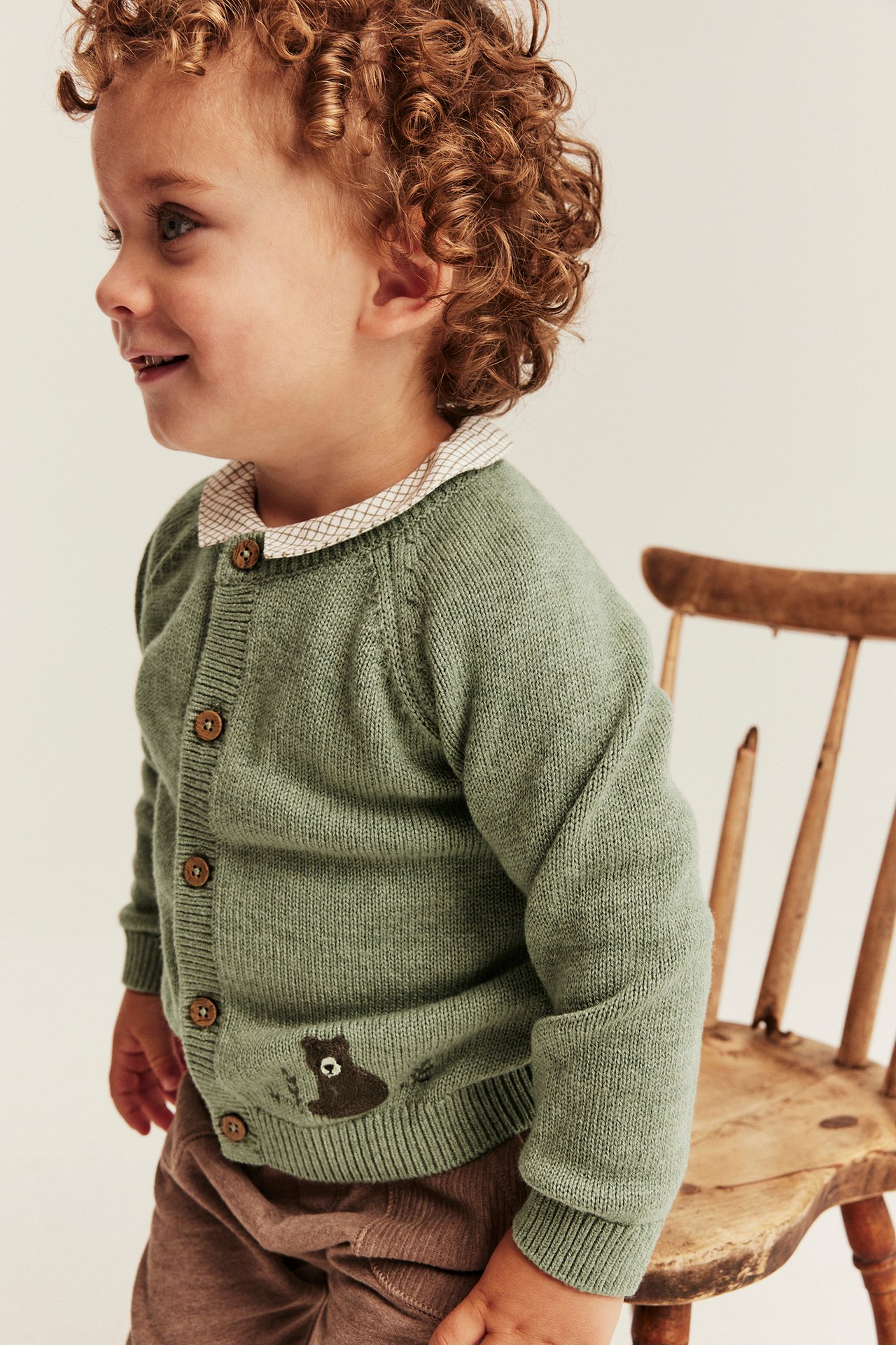 Grüne Strickjacke für Babys mit Knöpfen, Bärenstickerei und kariertem Hemd. Seitenansicht.