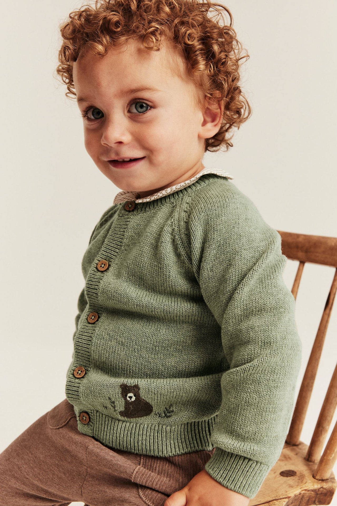 Vorderansicht eines Kindes in grünem Strickcardigan mit Bärenstickerei und brauner Hose.