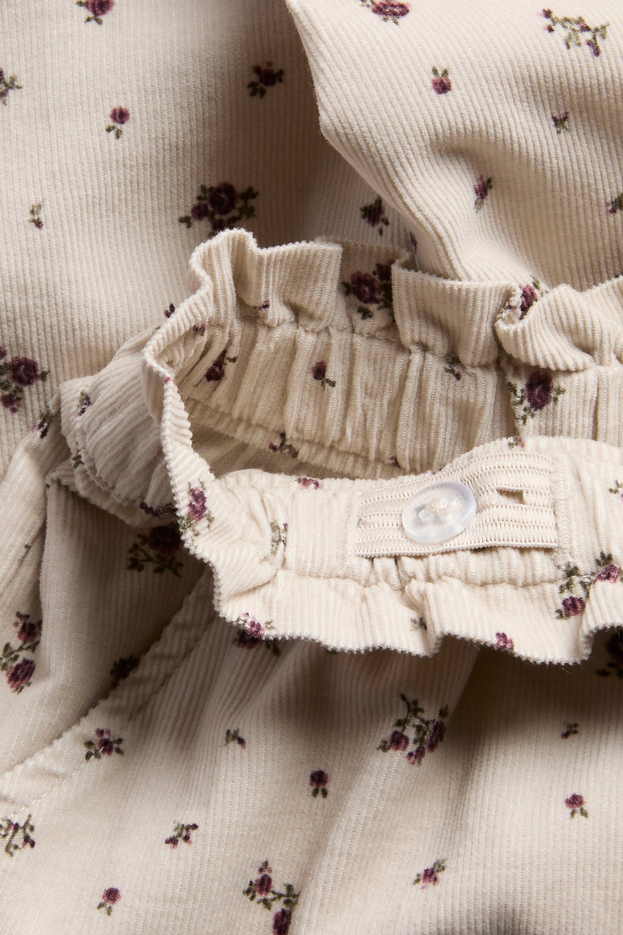 Nahaufnahme eines cremefarbenen Blumen-Cord-Kinderkleidungsstücks mit Rüschenbund und Knopf.