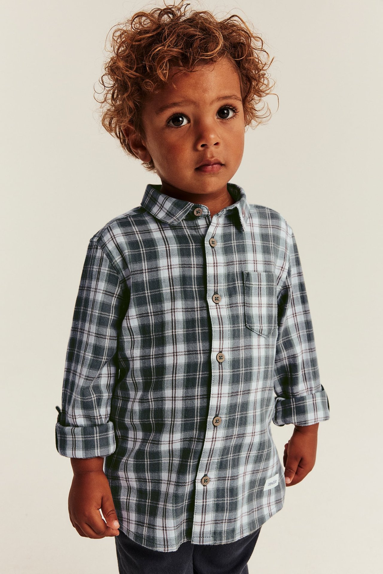 Front view of a kid wearing a dark gray and white plaid long-sleeve shirt with rolled-up sleeves and dark pants.