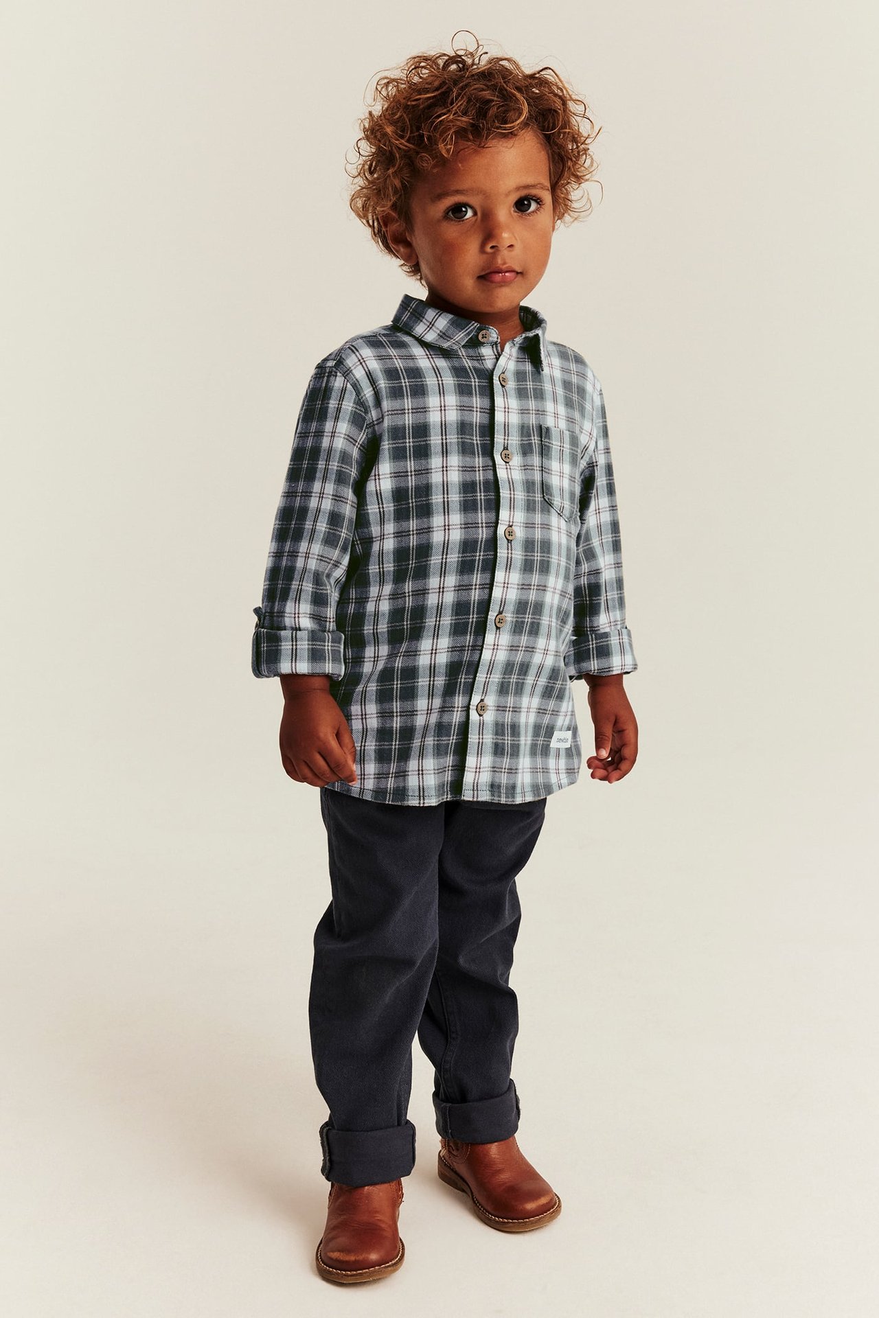 Front view of a kid wearing a dark gray plaid long-sleeved shirt, dark blue pants, and brown boots.