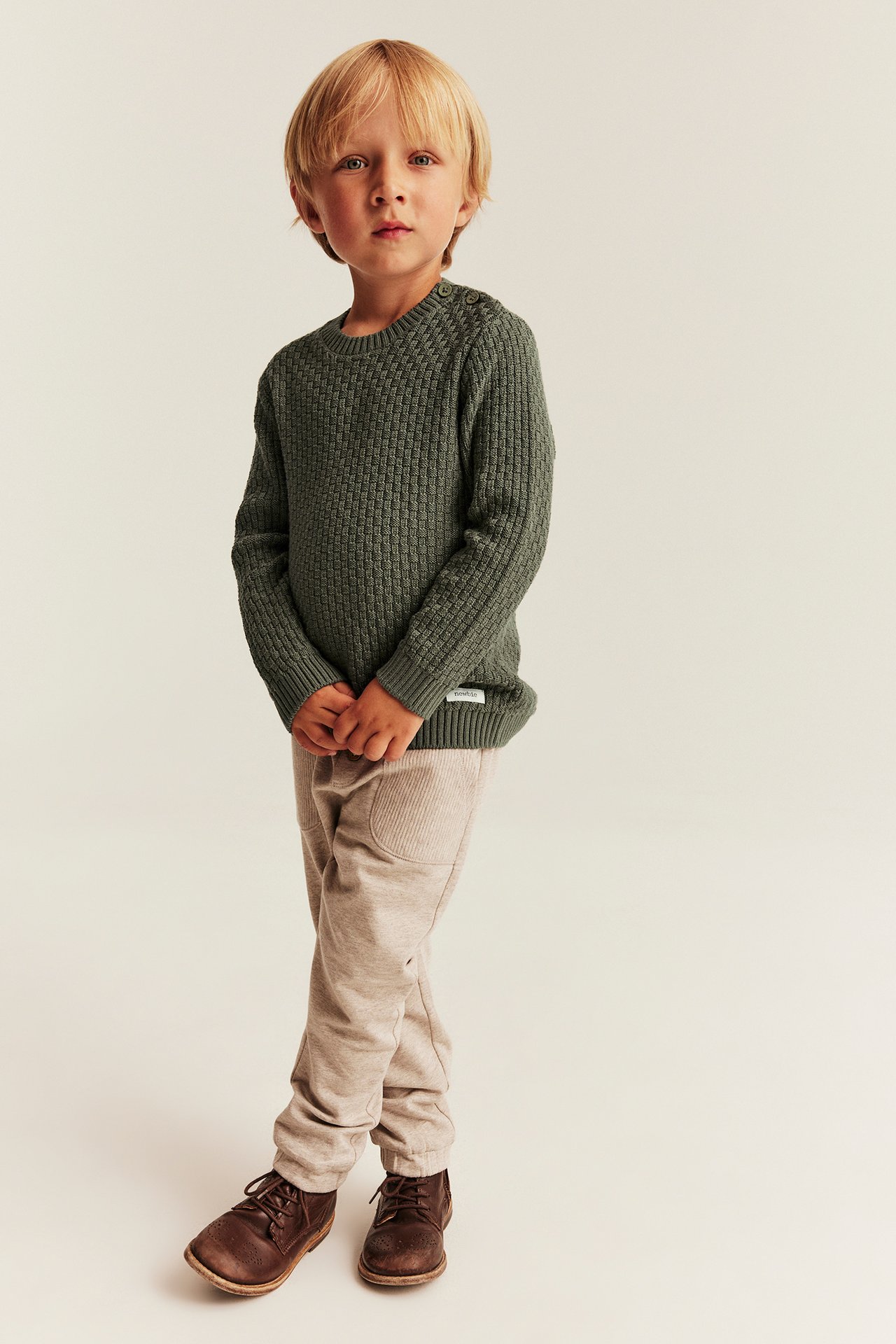 Vorderansicht: Kind in grünem Strickpullover, beiger Hose und braunen Schuhen.