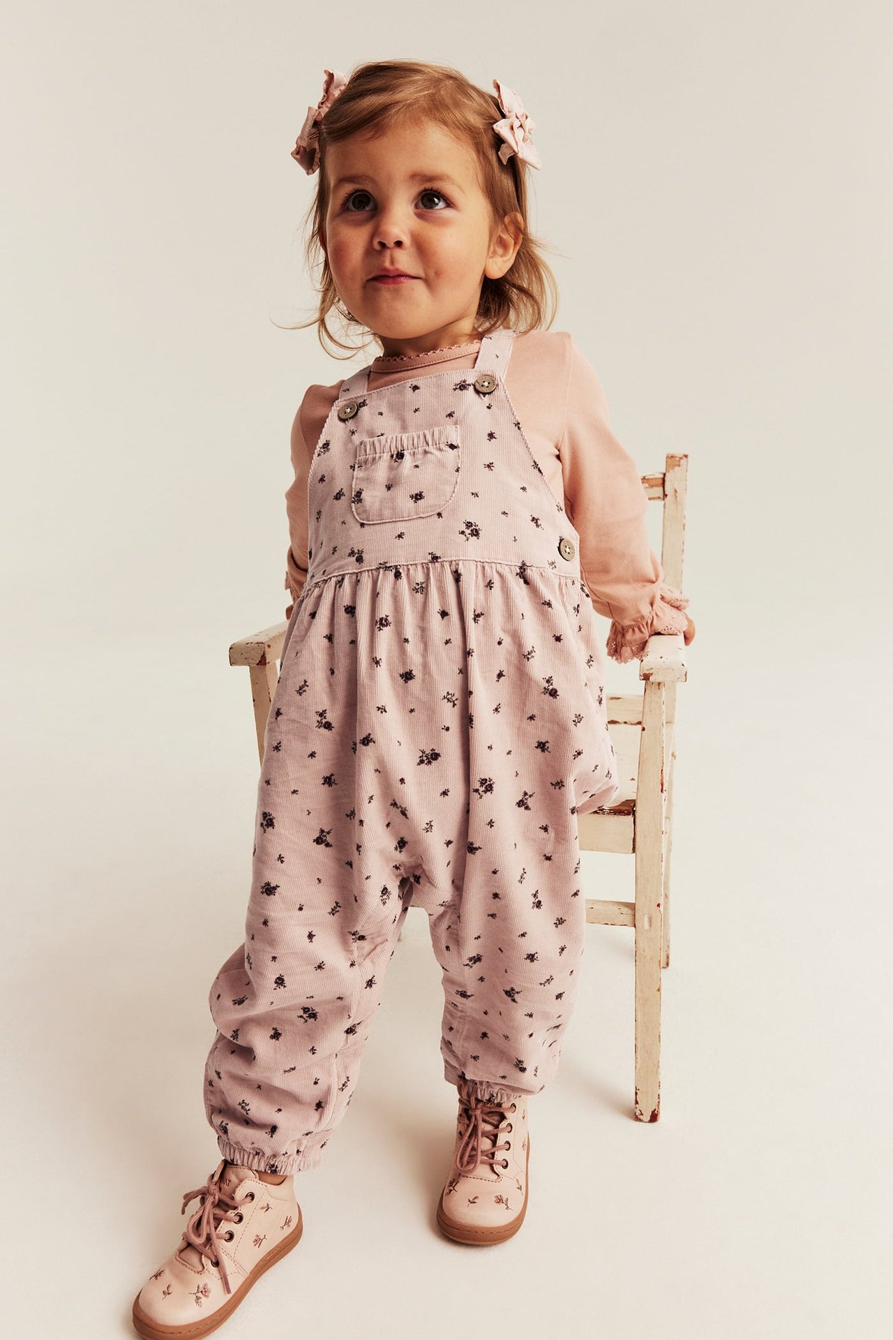 Vorderansicht, Kind in rosa Blumenoverall, rosa Shirt, rosa Schnürstiefeln und Haarschleifen.