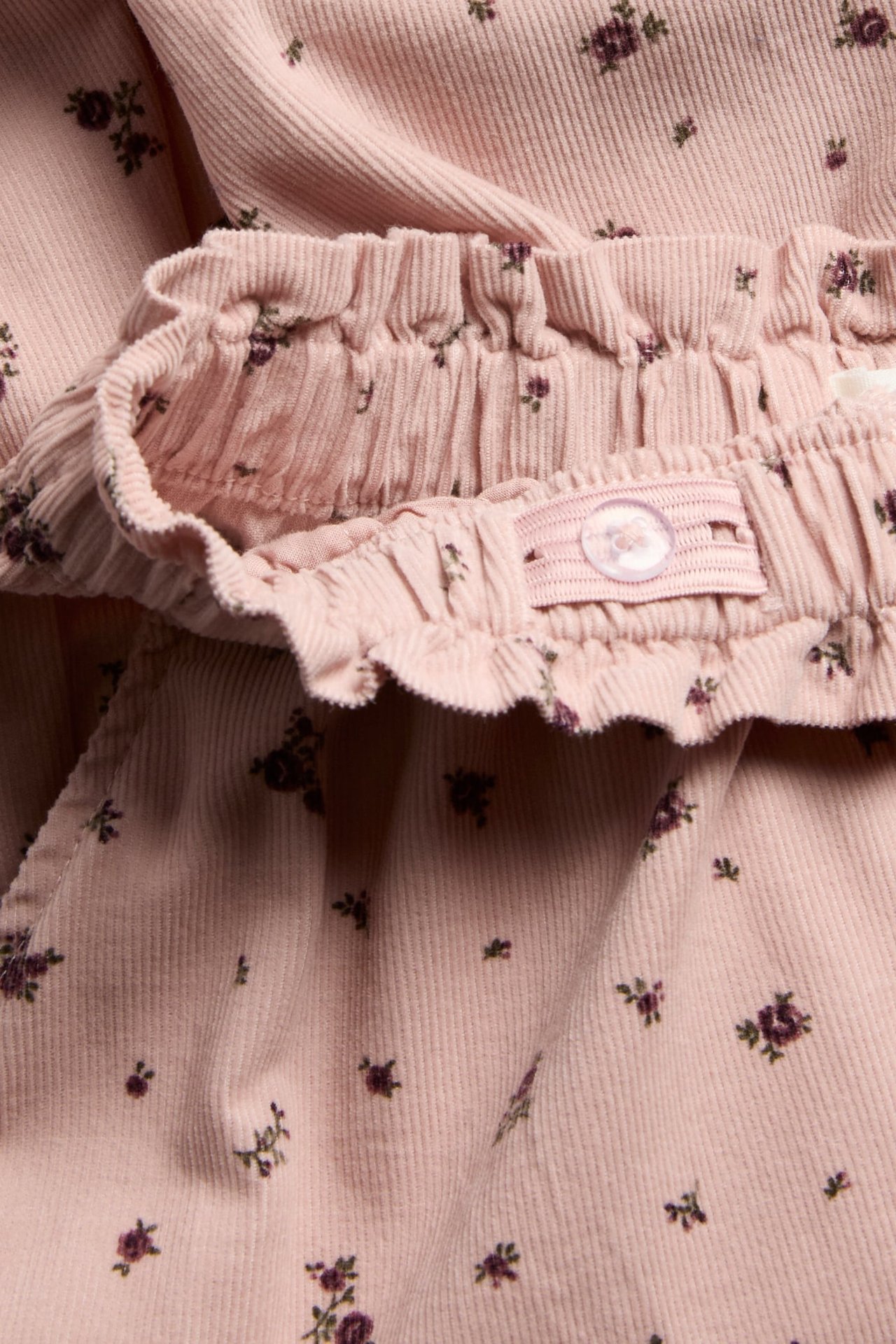 Nahaufnahme einer rosa geblümten Cordhose für Kinder mit elastischem Rüschenbund und verstellbarem Knopf.
