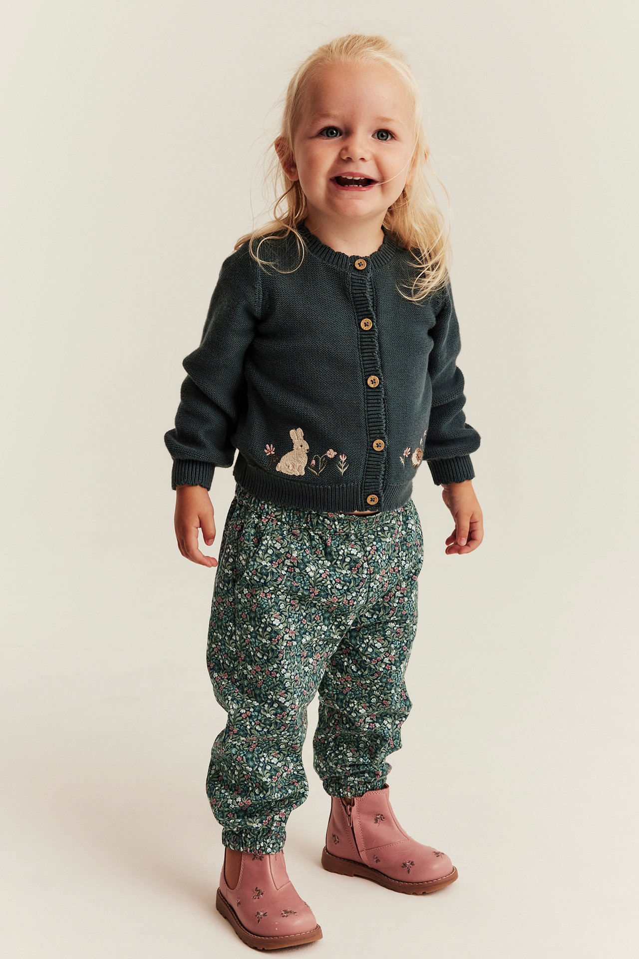 Front view of a kid wearing a dark green embroidered cardigan, floral print pants, and pink embroidered boots.