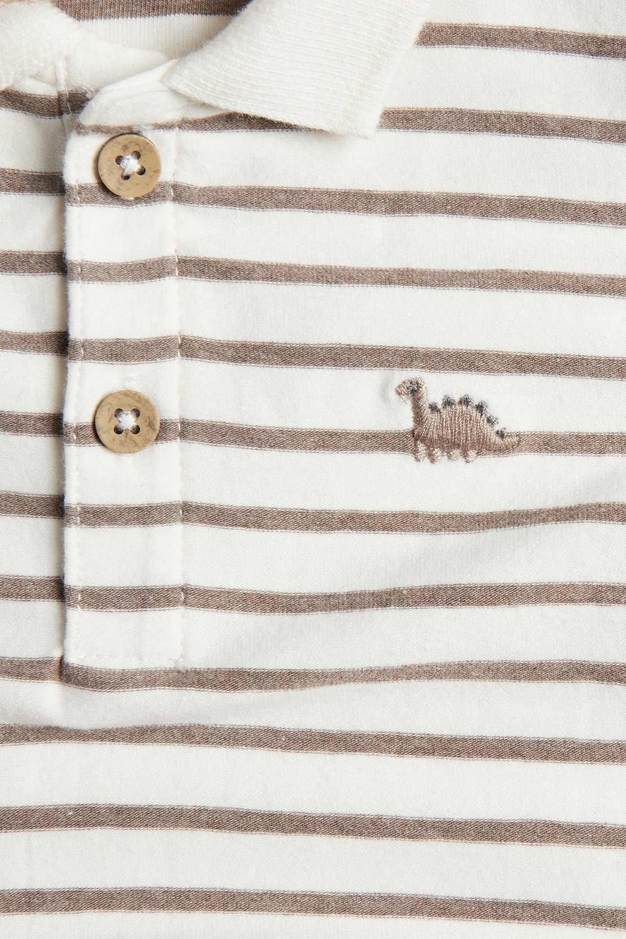 Close-up of a kid's white and brown striped polo shirt with two buttons and an embroidered dinosaur.