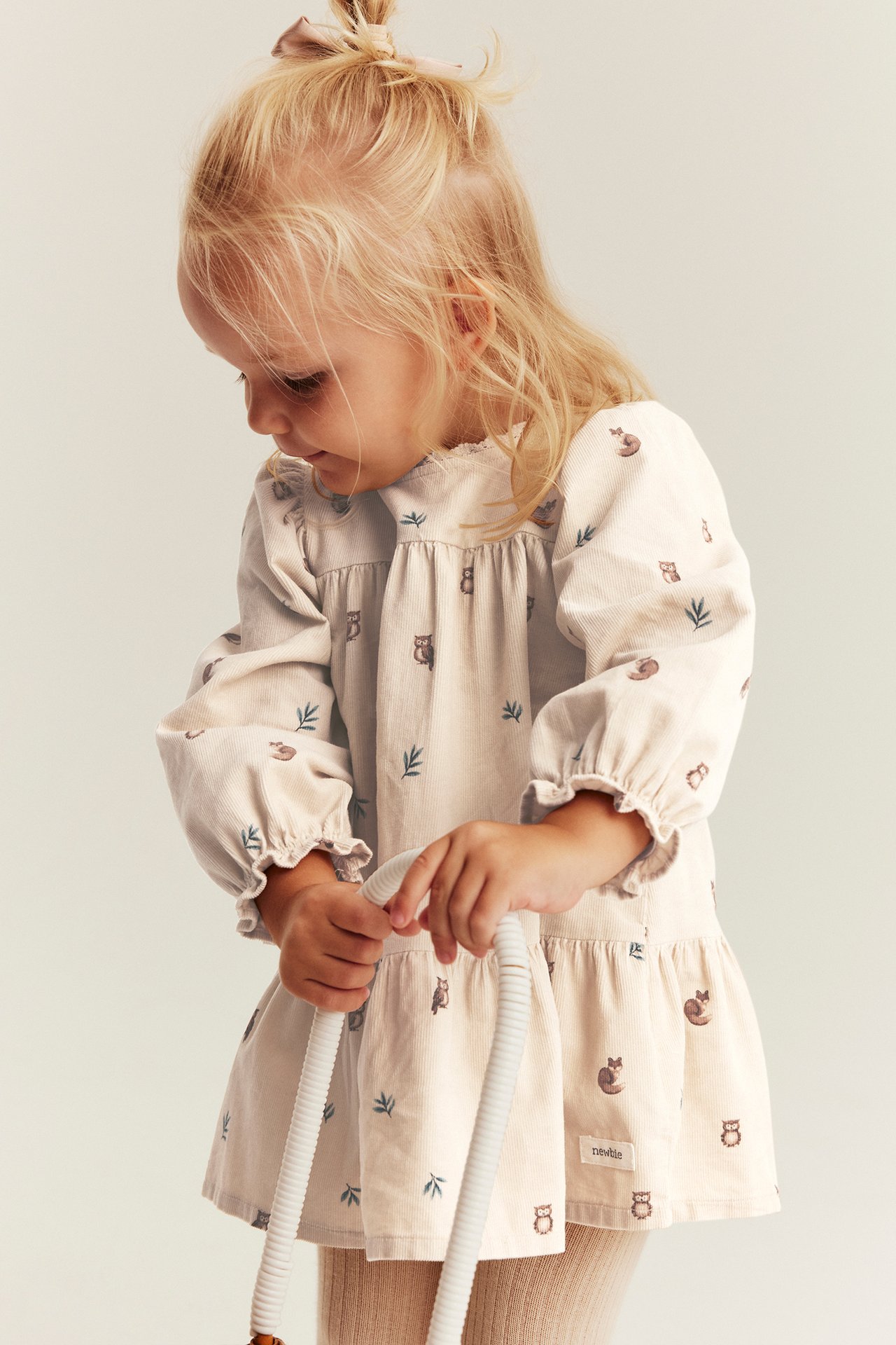 Vorderansicht Kind in cremefarbenem Cordkleid mit Waldtiermuster, hält weißes Spielzeug.