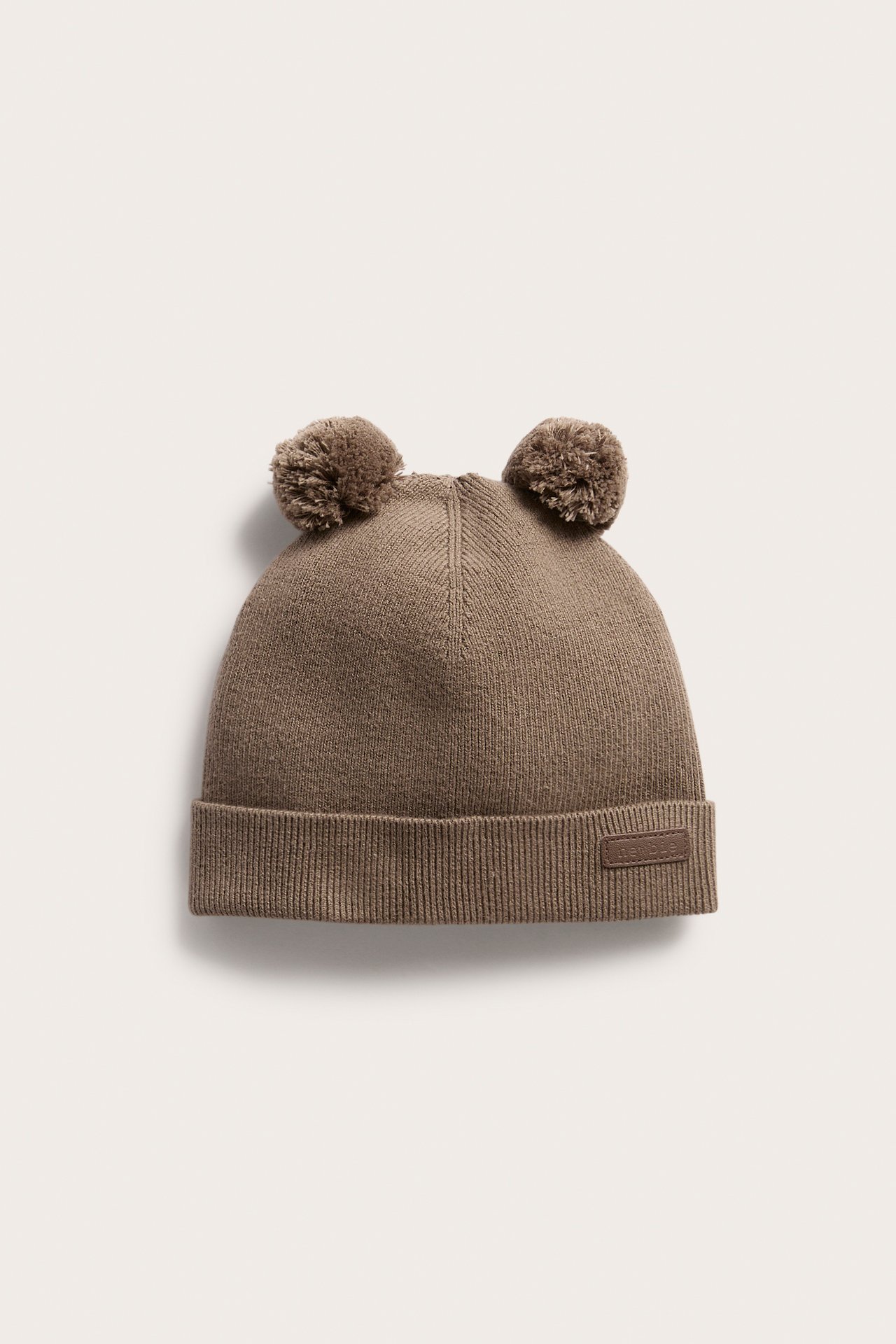 Front view of a brown knitted baby beanie with two pom-poms and a ribbed cuff.