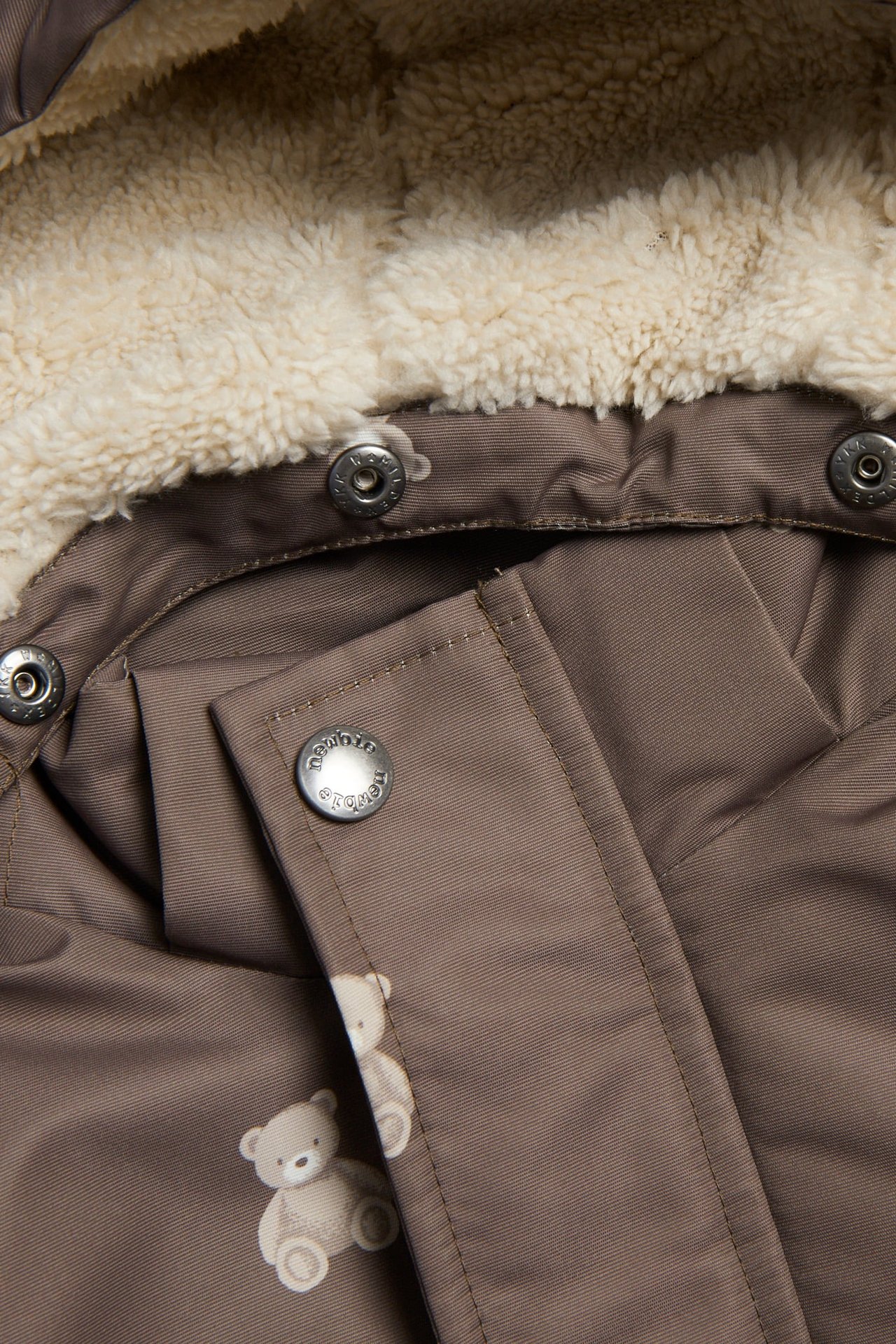 Nahaufnahme einer braunen Babyjacke mit cremefarbener Kapuze aus Kunstfell, Druckknöpfen und Bärchenmuster.