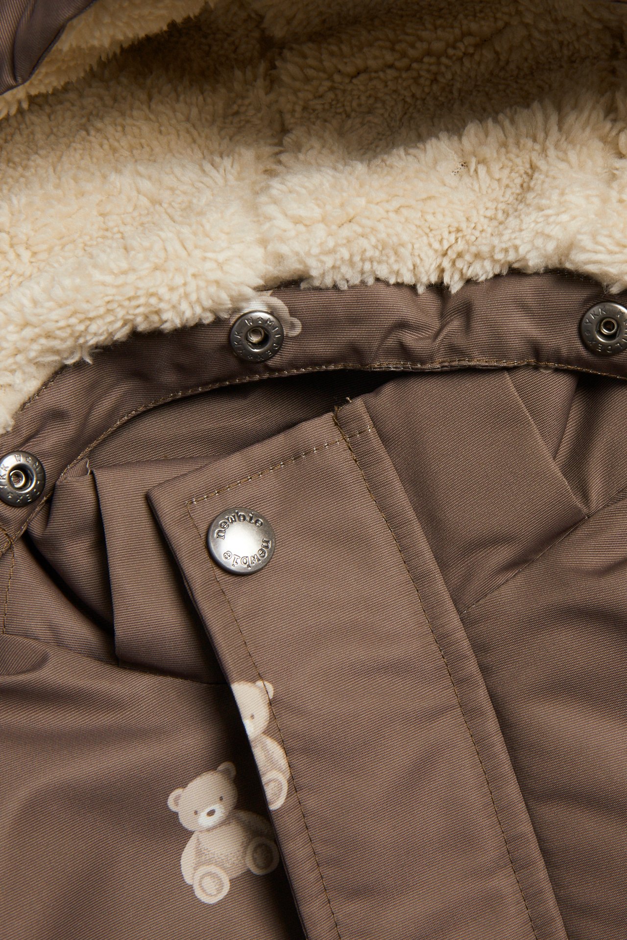 Nahaufnahme brauner Kinderjacke mit cremefarbenen Teddybären, cremefarbenem Futter und silbernen Druckknöpfen.