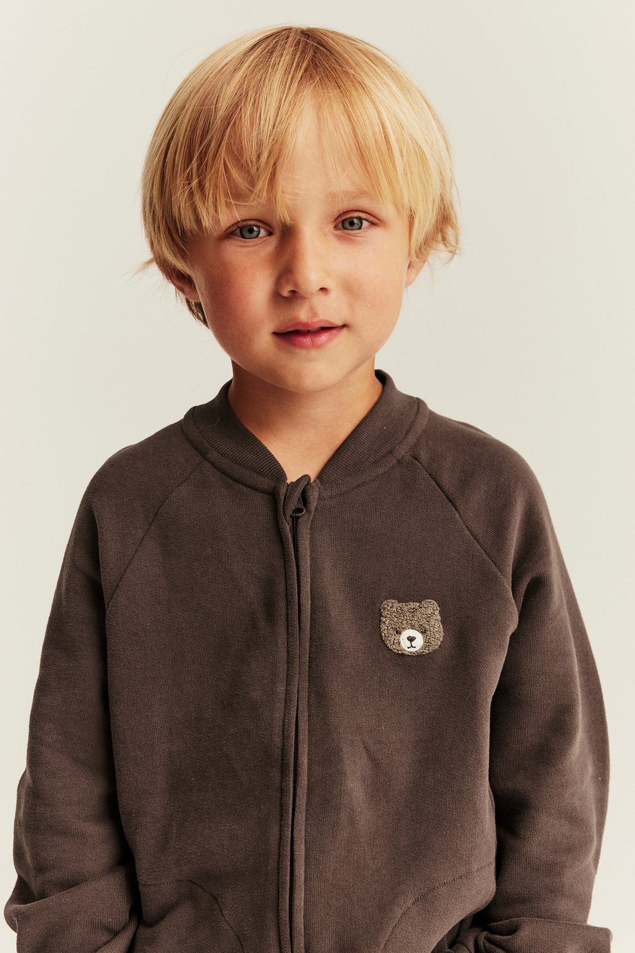 Vorderansicht: Kind in dunkelbraunem Reißverschluss-Sweatshirt mit Teddybär-Patch.