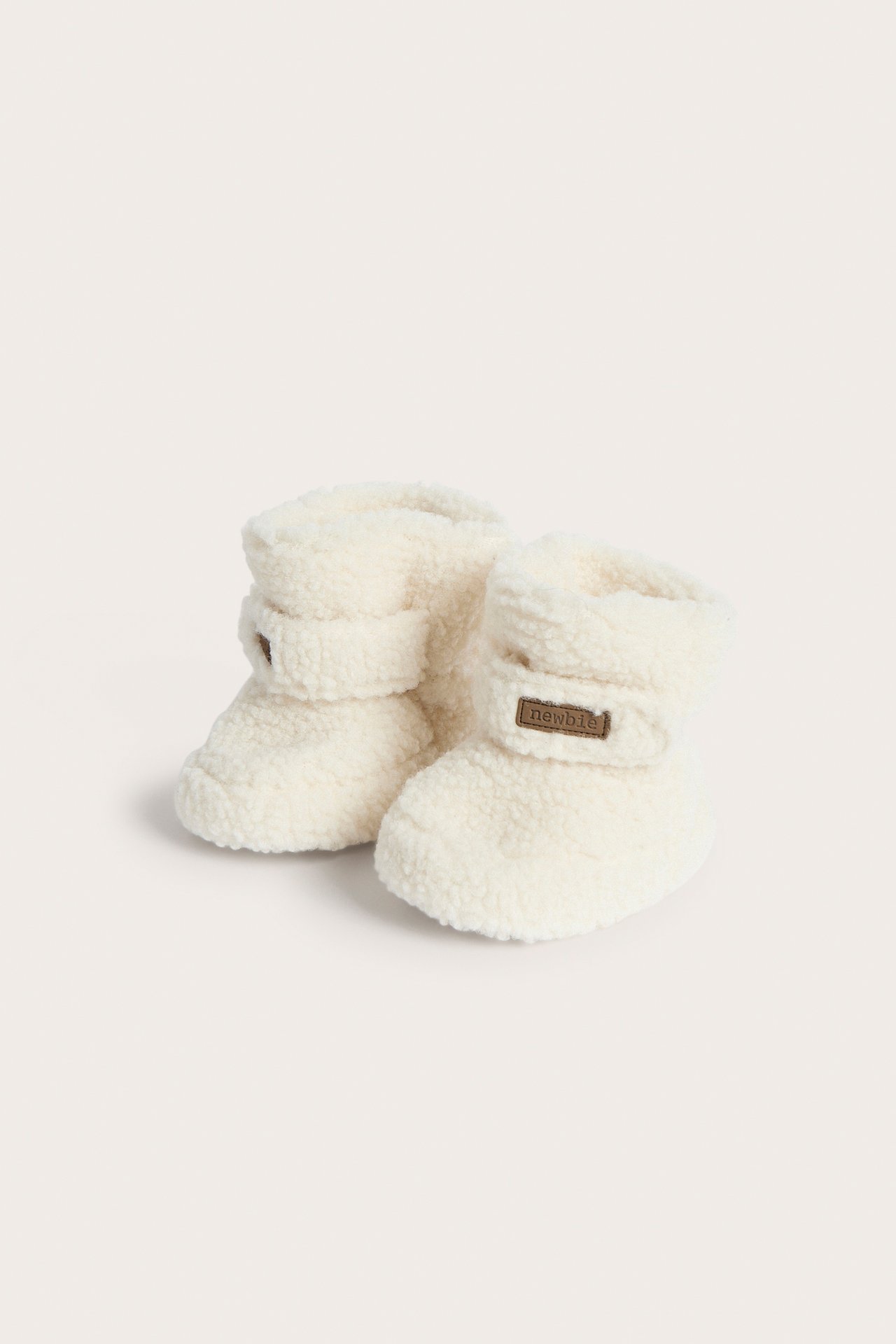 Front-side view of a pair of cream teddy baby booties with a small brown 'newbie' label.