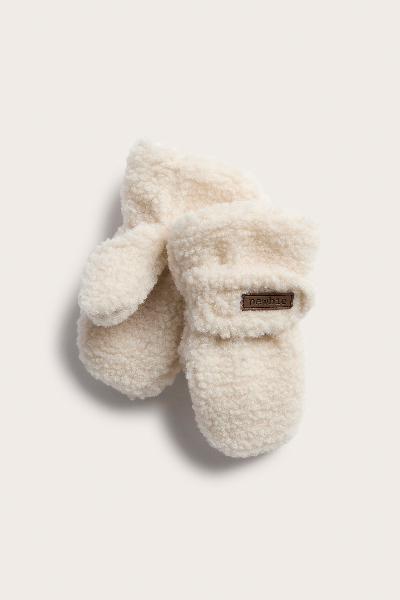 Top-down view of a pair of cream fluffy baby mittens with a brown label.
