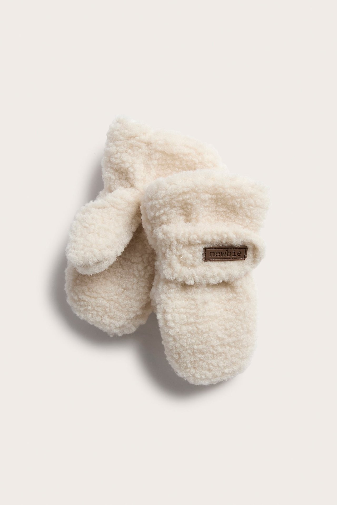 Draufsicht auf ein Paar cremefarbene flauschige Babyfäustlinge mit braunem Etikett.