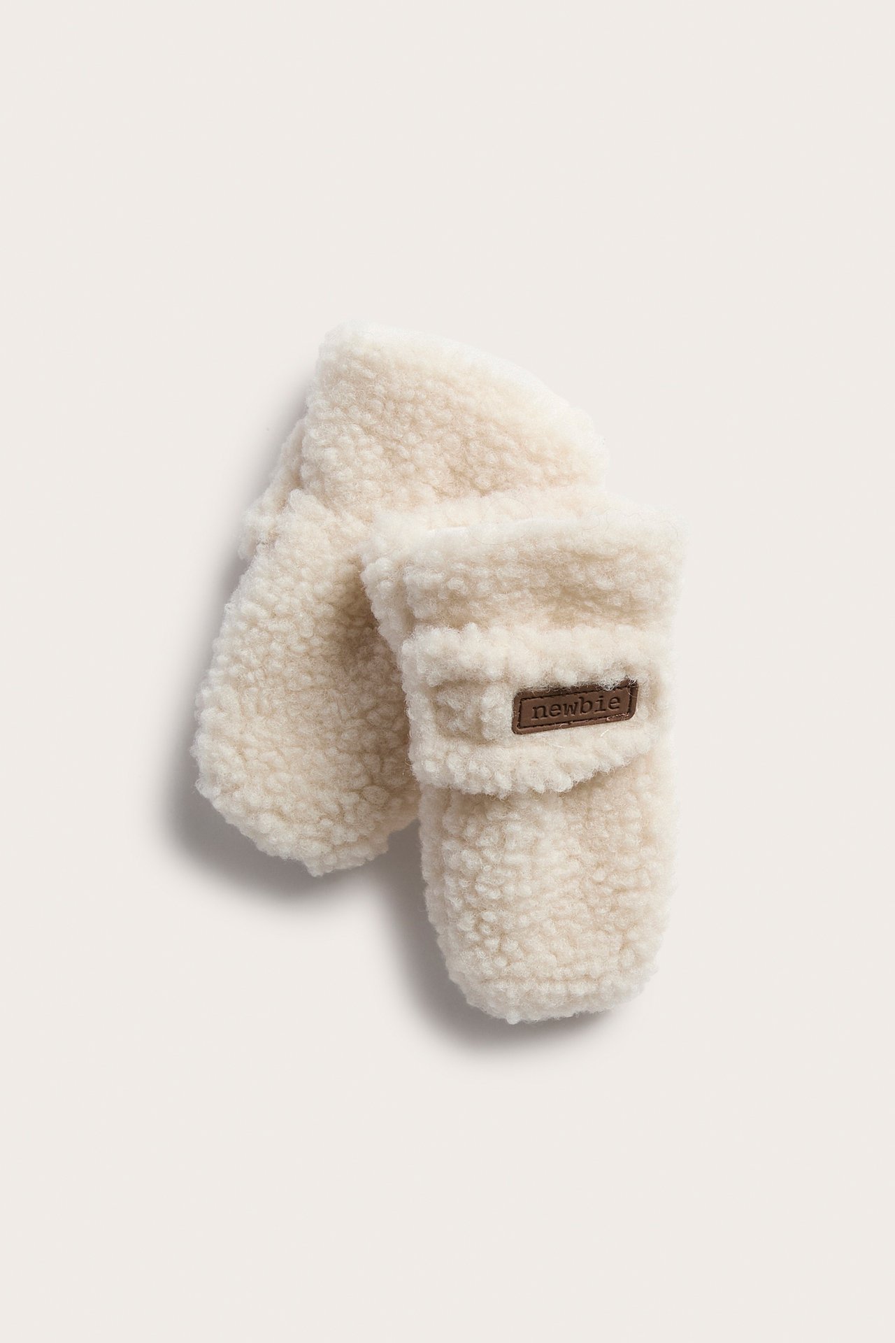 Top view of a pair of cream fluffy baby mittens with a brown label.
