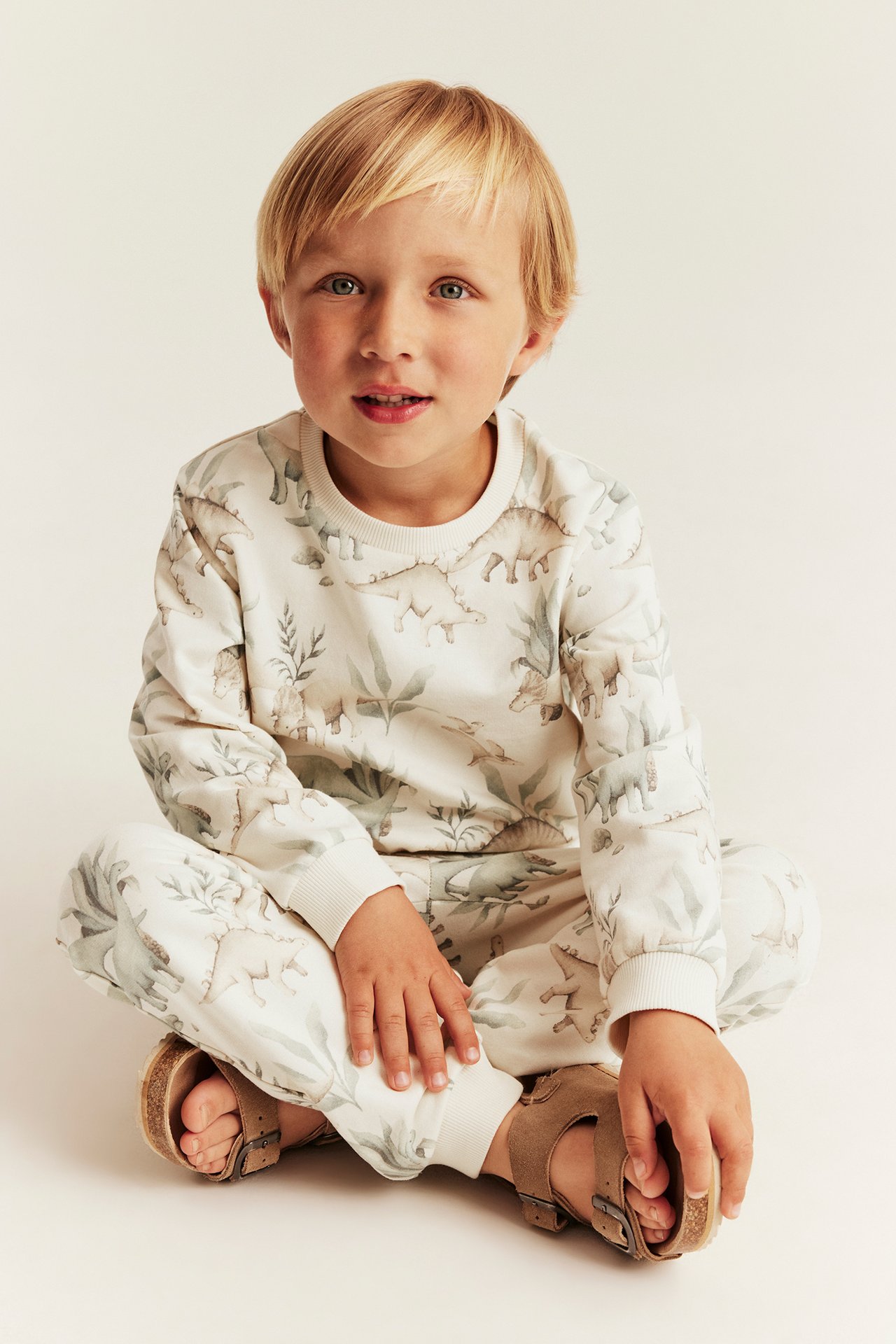 Vorderansicht, Kind trägt cremefarbenes Sweatshirt mit Dinosauriermuster und passende Jogginghose mit beigen Sandalen.