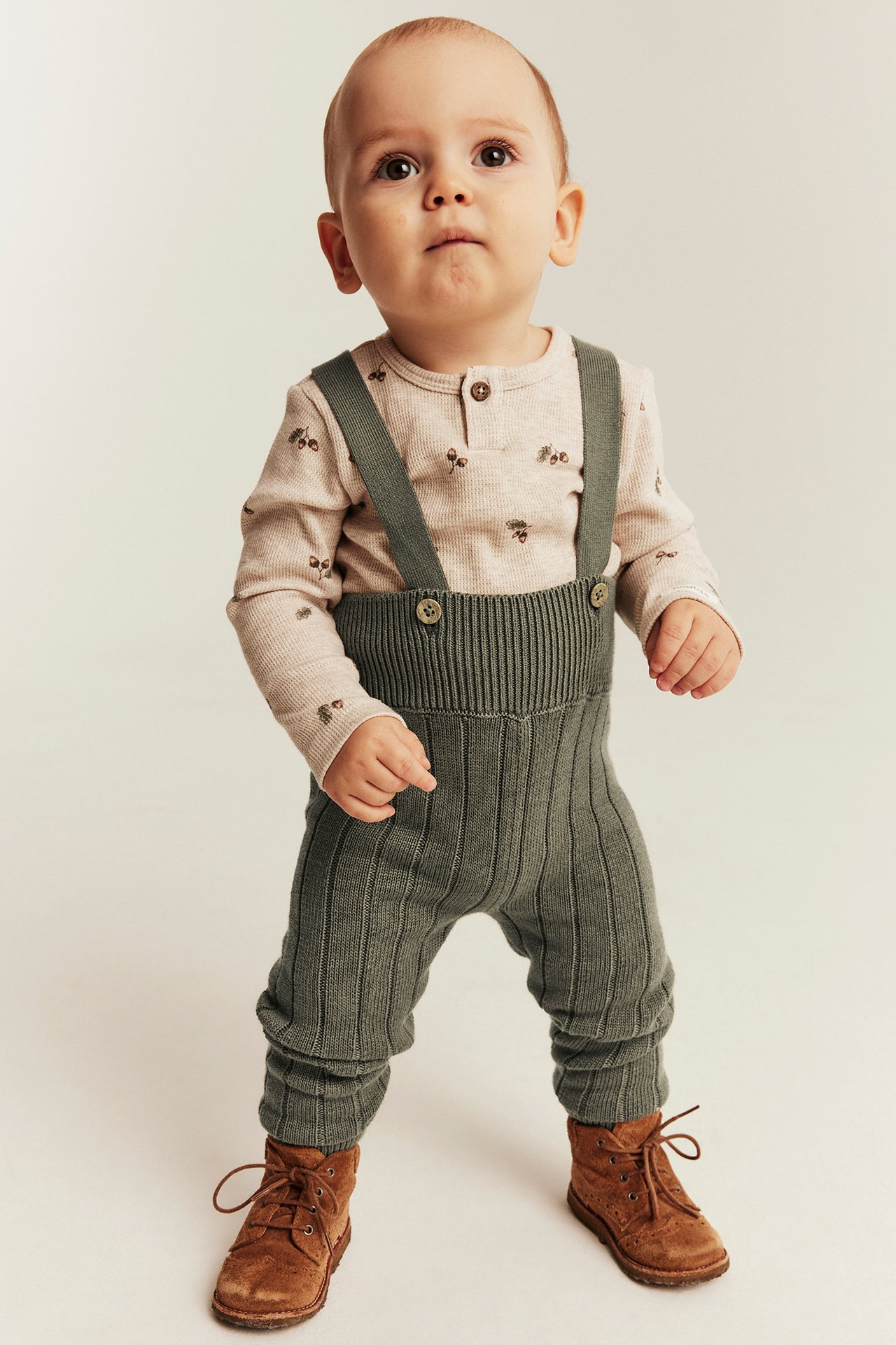 Green ribbed knit overalls, beige patterned long-sleeve top, and brown boots on baby, front view.