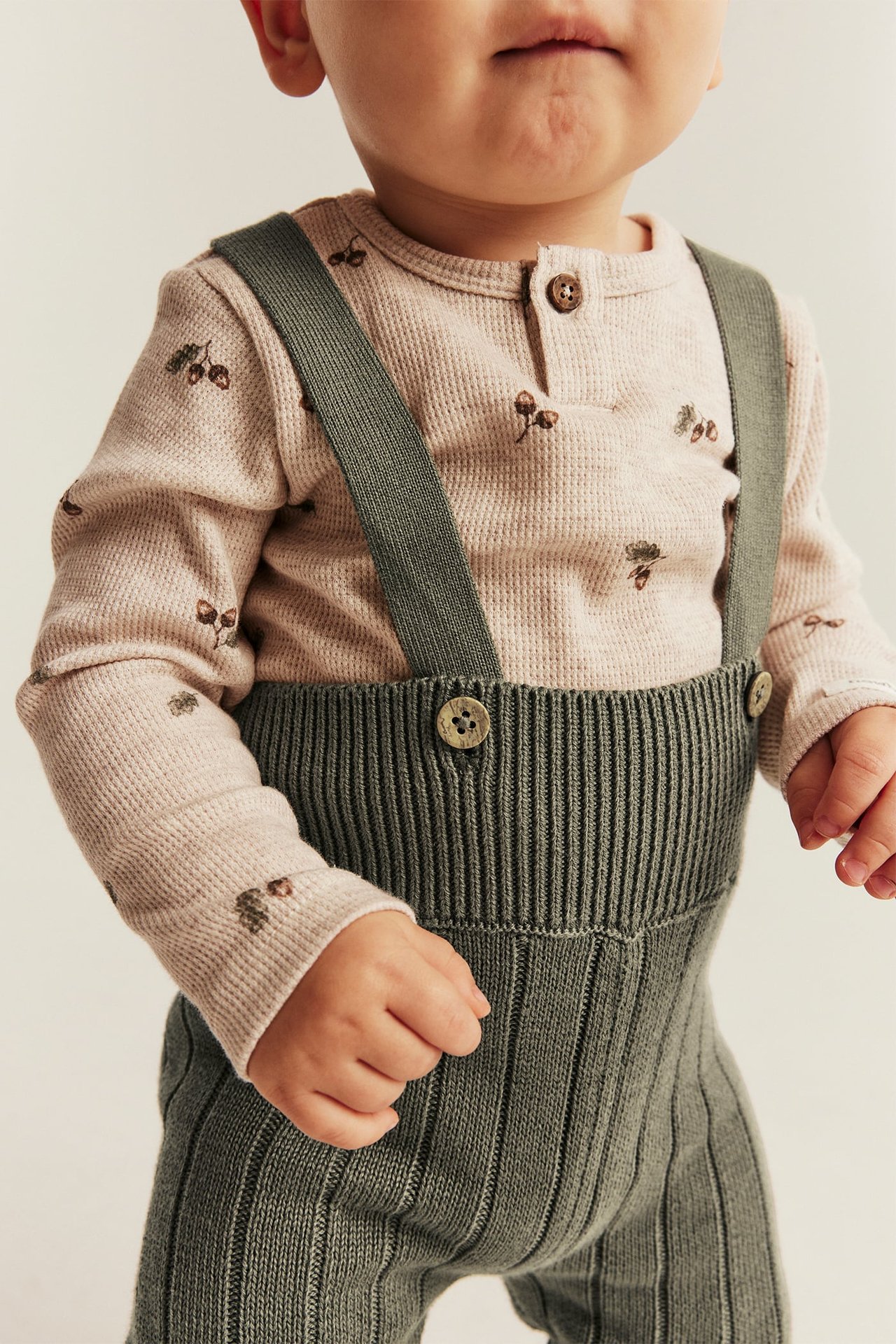 Vorderansicht eines Babys in grünen gerippten Stricklatzhosen und einem rosa Waffel-Oberteil mit Eichelprint.