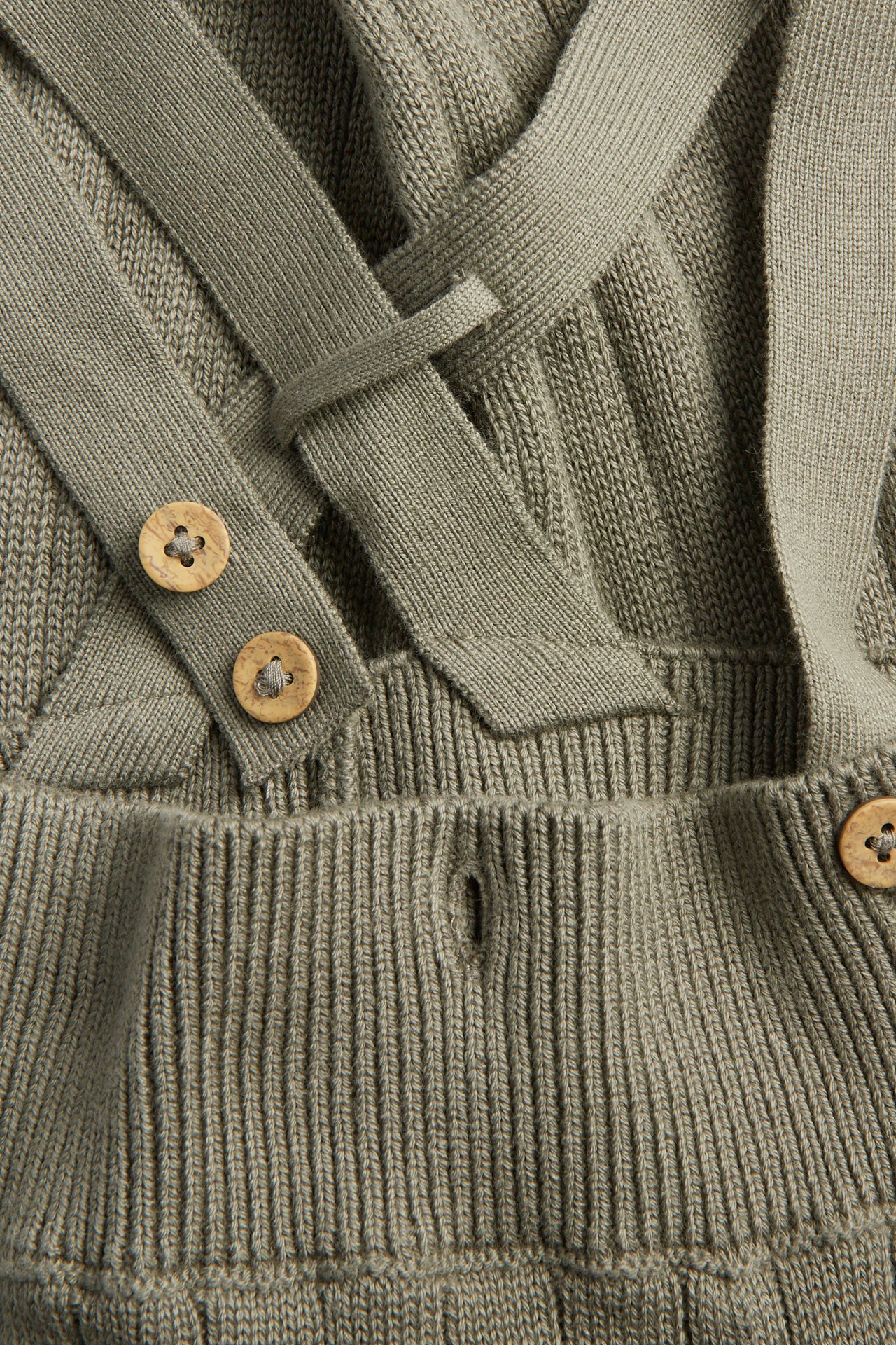 Close-up of a khaki knitted baby romper with criss-cross straps and wooden buttons.