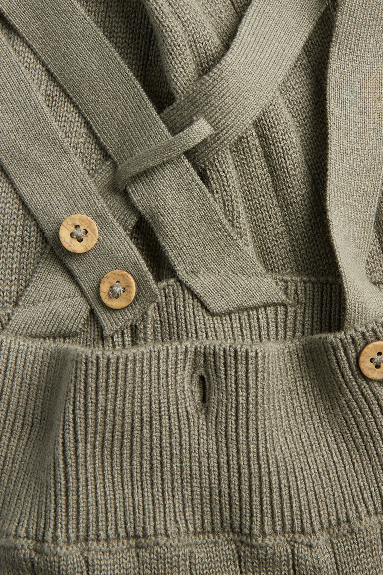 Nahaufnahme eines khakifarbenen gestrickten Baby-Stramplers mit überkreuzten Trägern und Holzknöpfen.