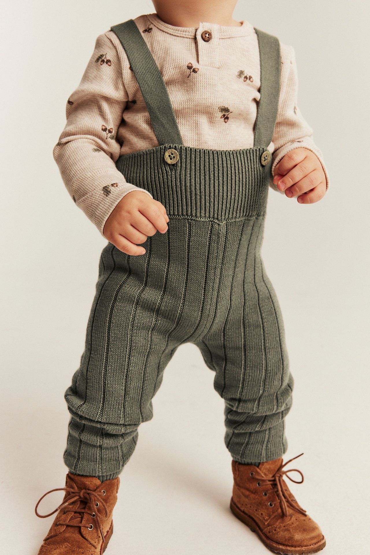 Front view: baby in green ribbed knit overalls, a patterned long-sleeve top, and brown lace-up boots.