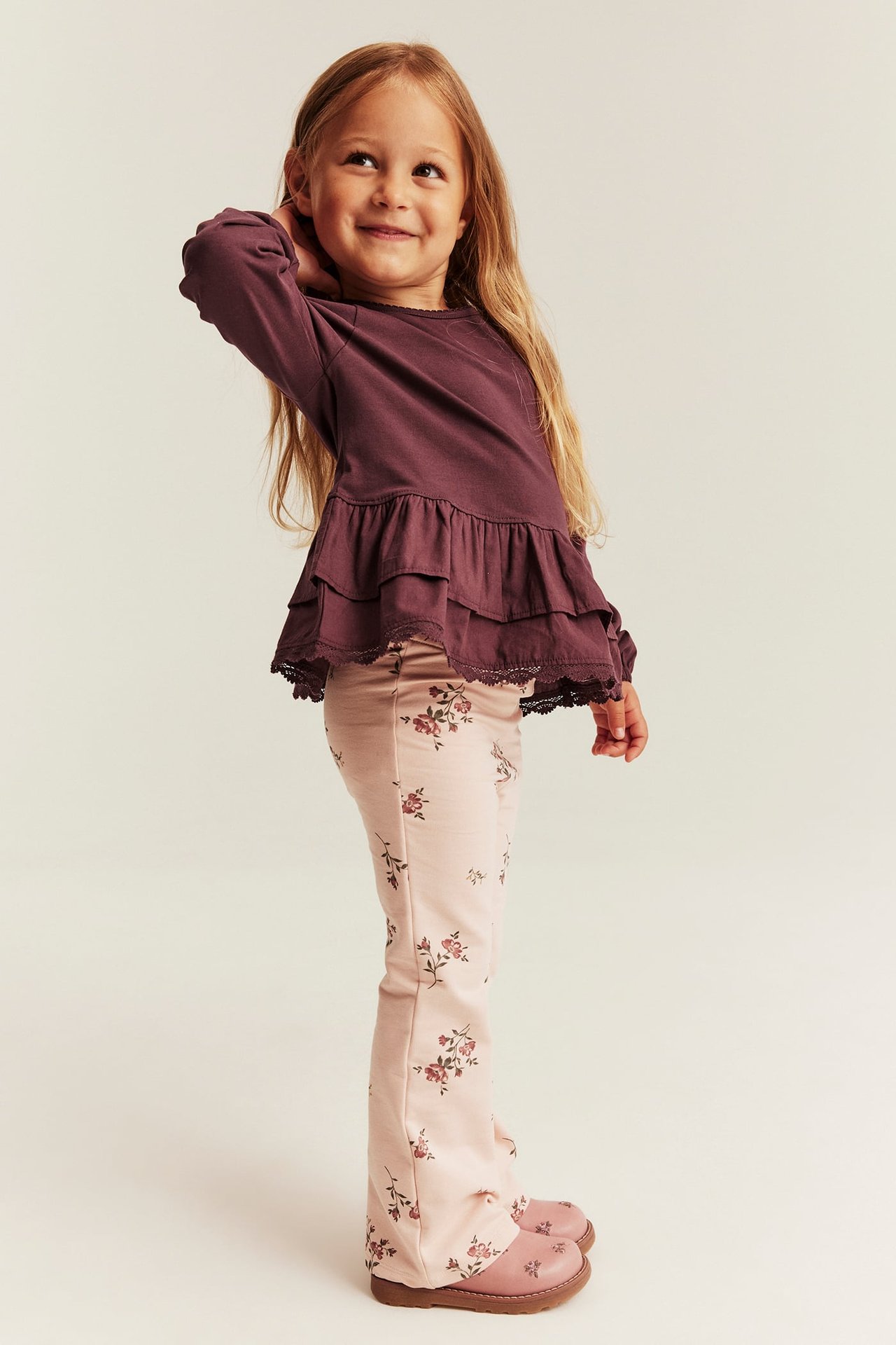 Seitenansicht von Kind in pflaumenfarbenem Langarm-Peplum-Oberteil, hellrosa Blumen-Schlaghose und pinken Schuhen.