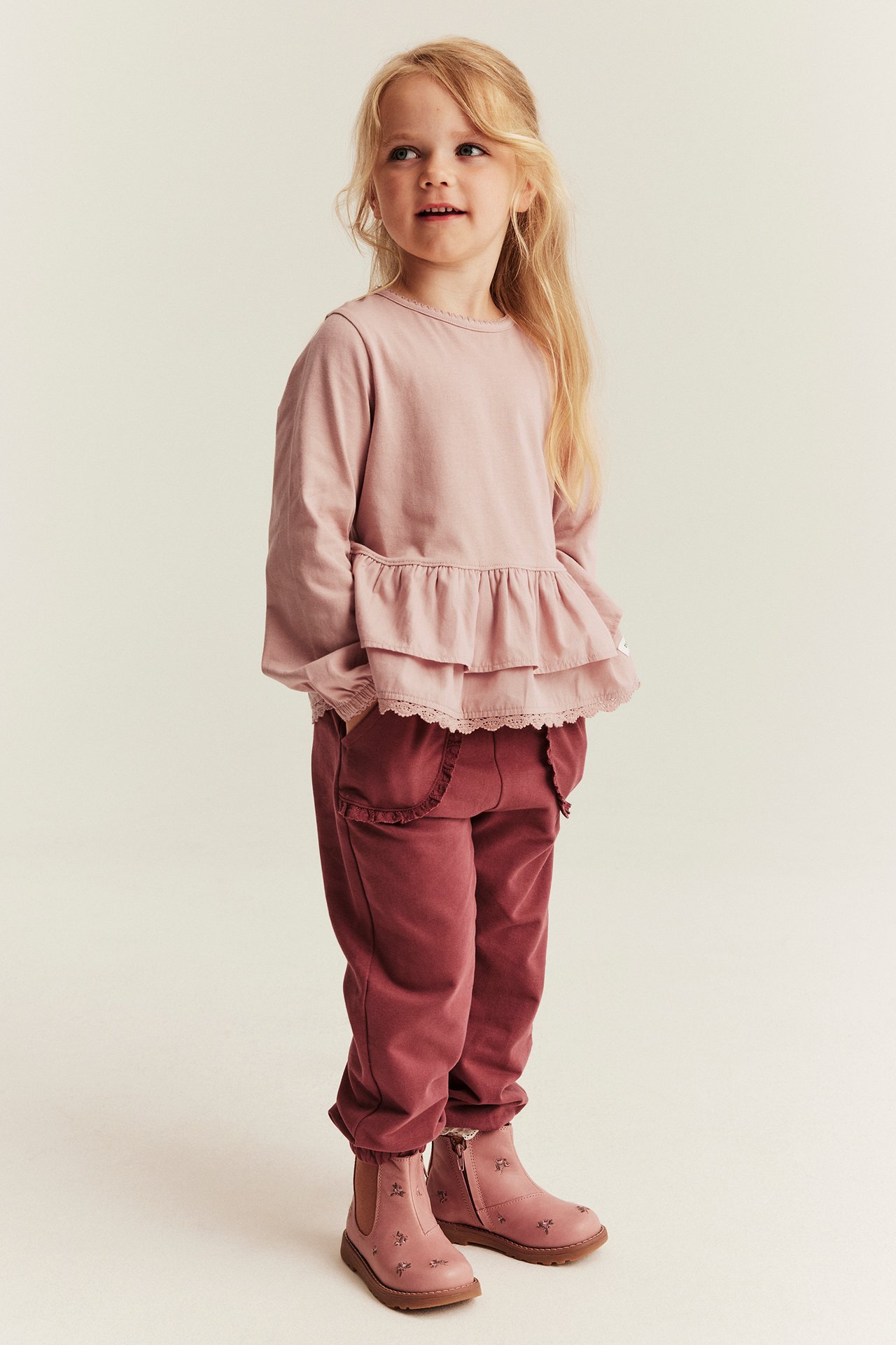 Frontansicht: Kind trägt hellrosa Rüschen-Langarmtop, weinrote Jogginghose, rosa bestickte Stiefeletten.