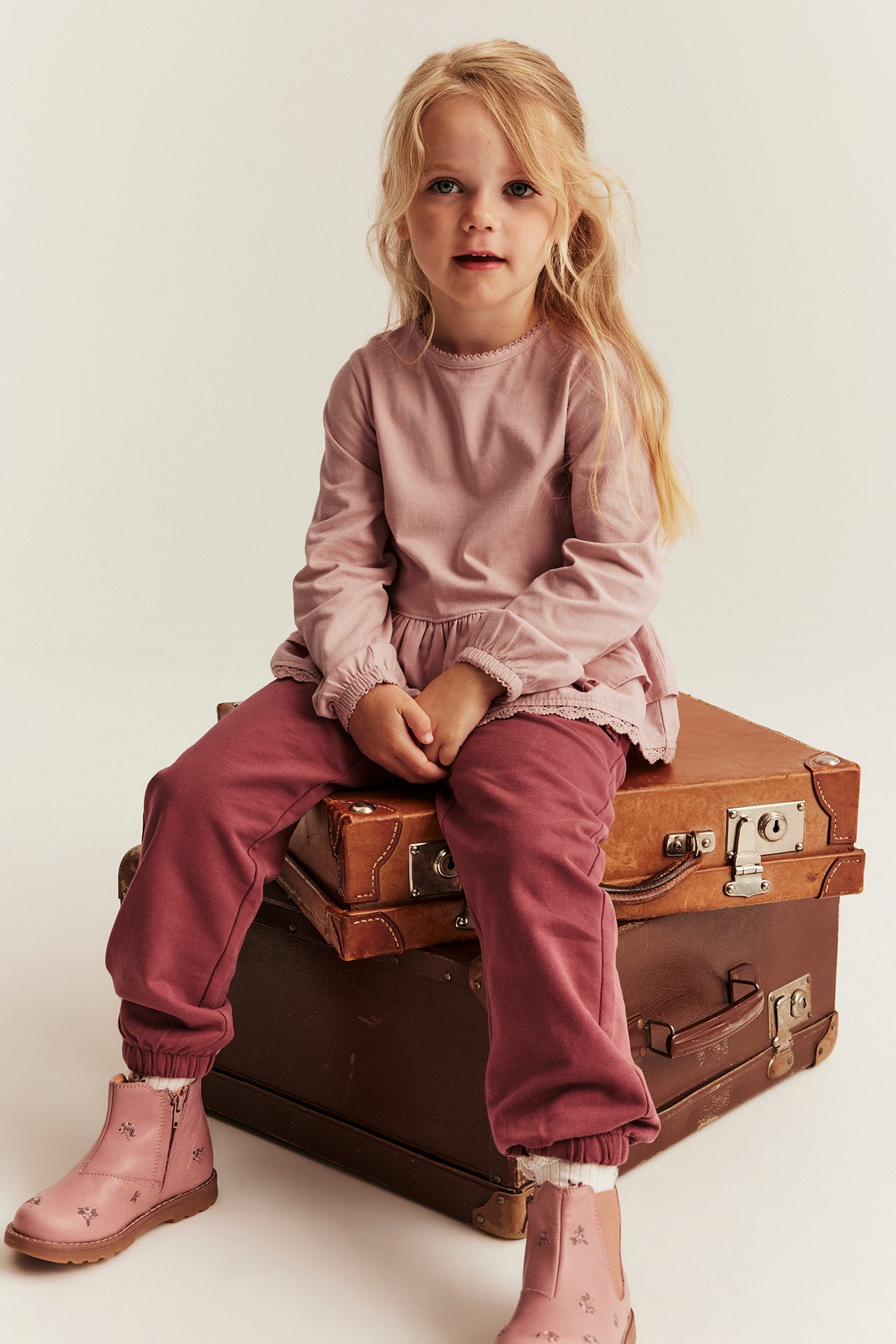 Front view of a kid wearing a pink long-sleeved peplum top, dark pink joggers, and pink embroidered ankle boots.