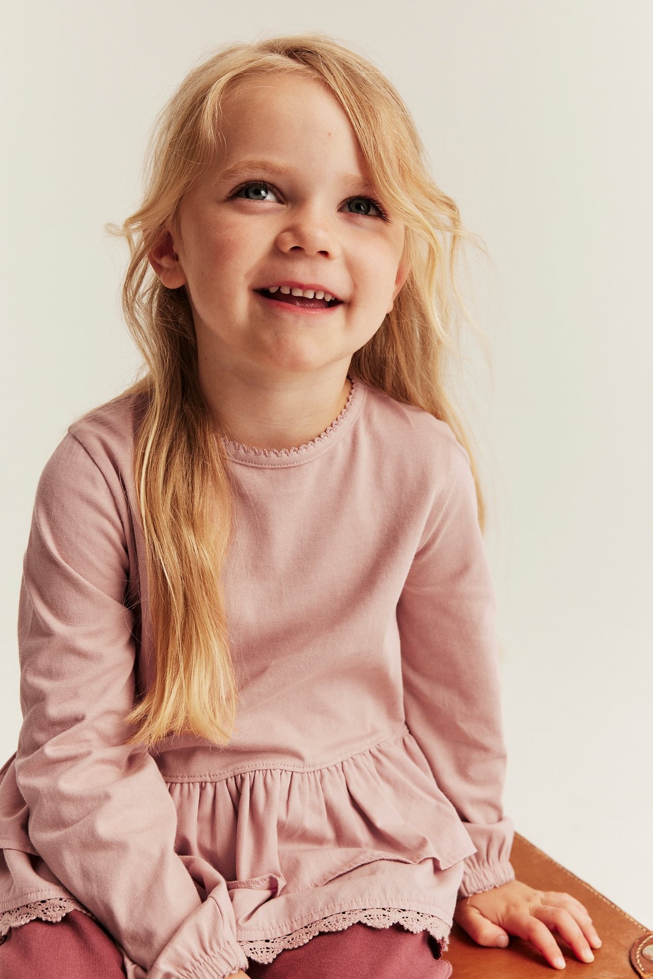 Kid wears dusty pink long-sleeved ruffle top with lace trim and dark pink leggings, front view.