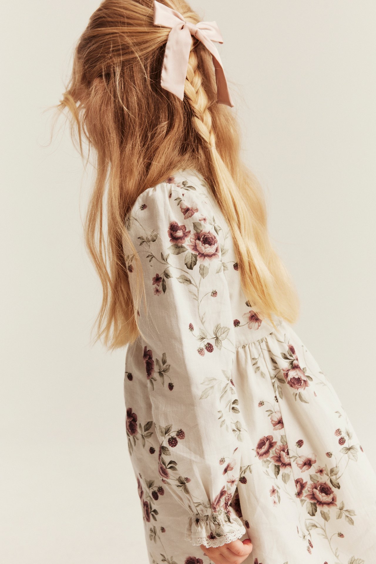 Seitenansicht: Kind trägt cremefarbenes Blumenkleid mit rosa Haarschleife.