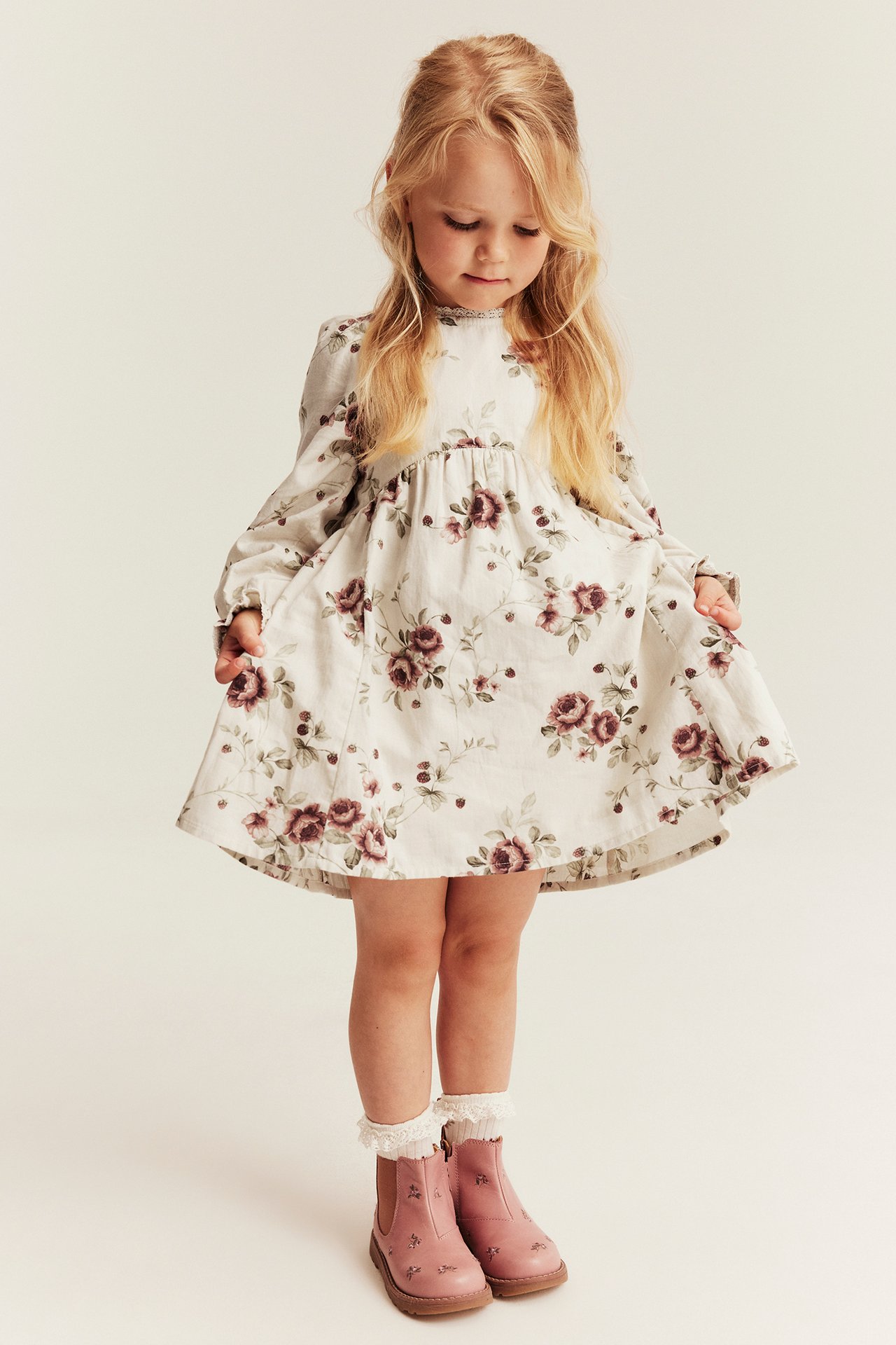 Vorderansicht eines Kindes in langärmeligem Blumenkleid, rosa Stiefeln und weißen Spitzensocken.