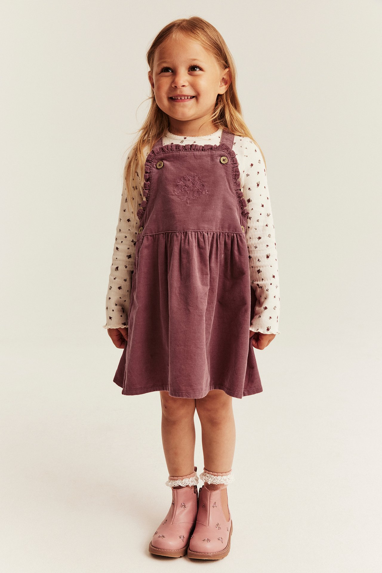 Front view: Kid in purple corduroy overall dress with embroidery, floral top, pink boots, and lace socks.