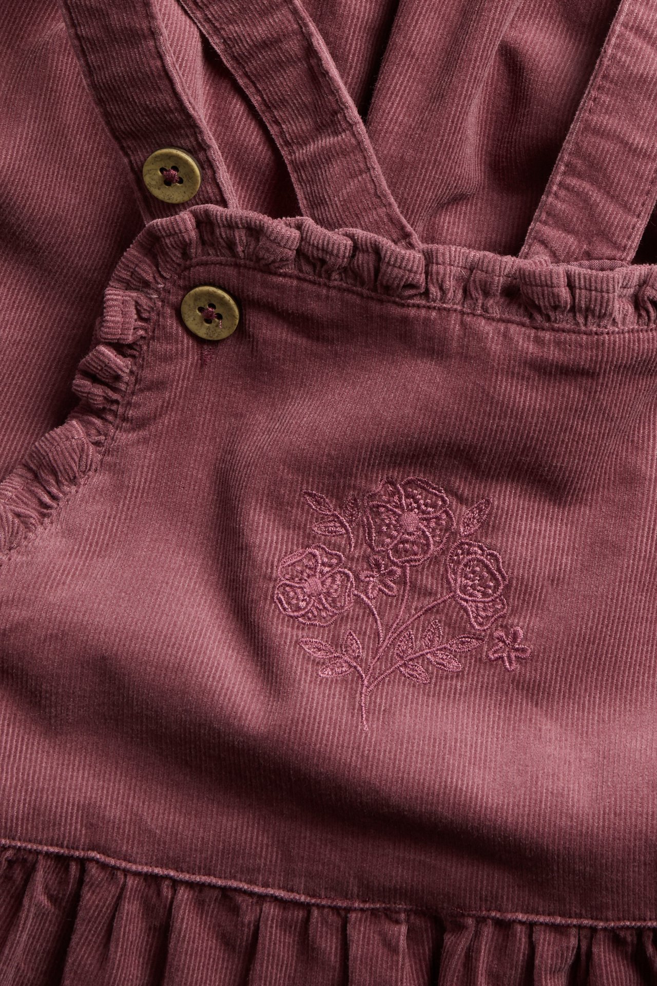 Close-up of a plum corduroy overall dress with strap buttons, ruffles, and floral embroidery.