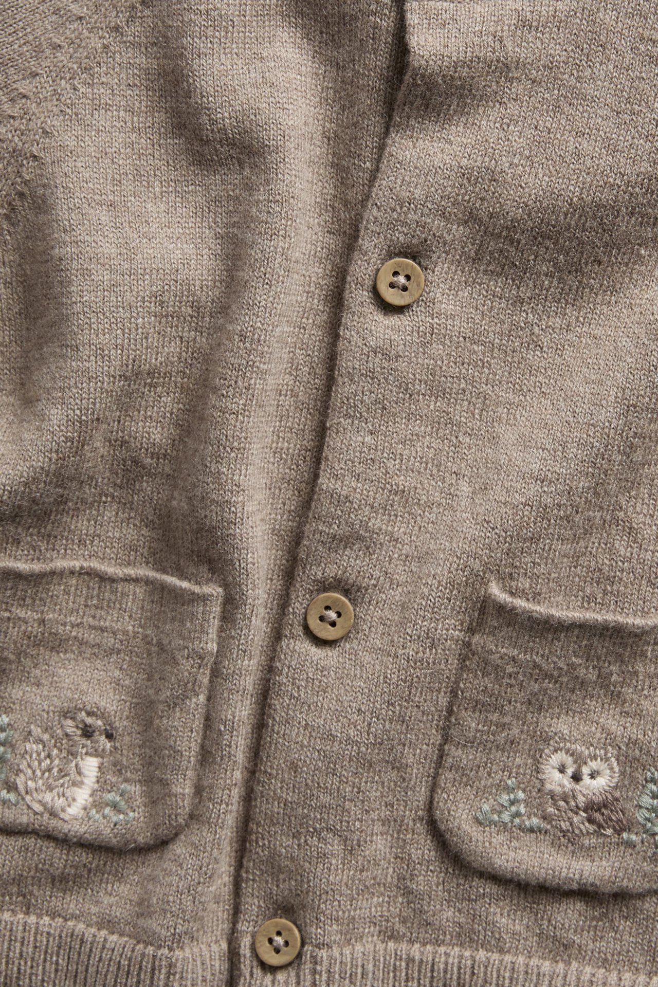 Nahaufnahme einer hellbraunen Strickjacke mit Knöpfen vorn und zwei Taschen mit Eulenstickerei.