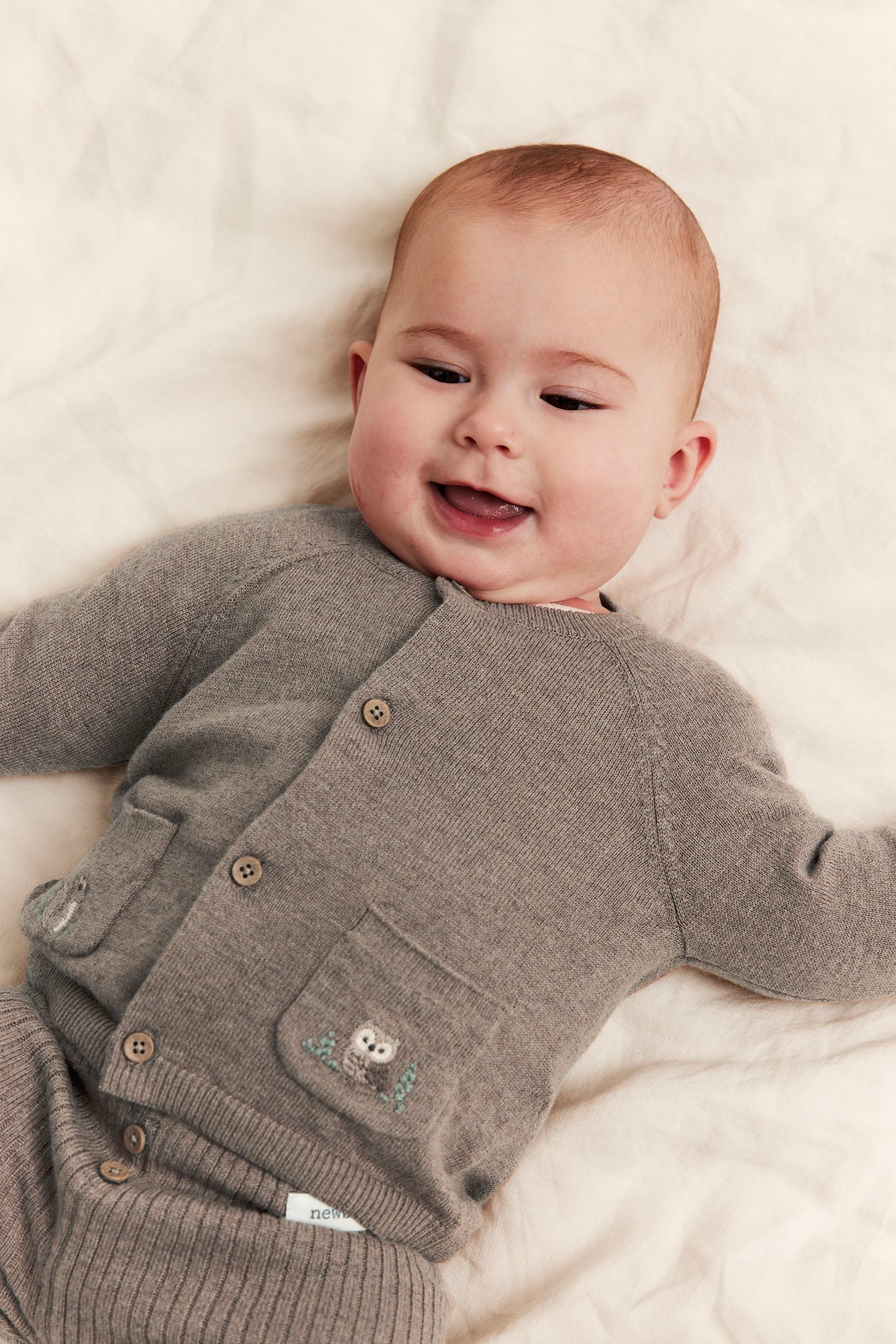 Vorderansicht eines Babys in grauer Knopfstrickjacke mit Eulenstickerei und passender gerippter Hose.