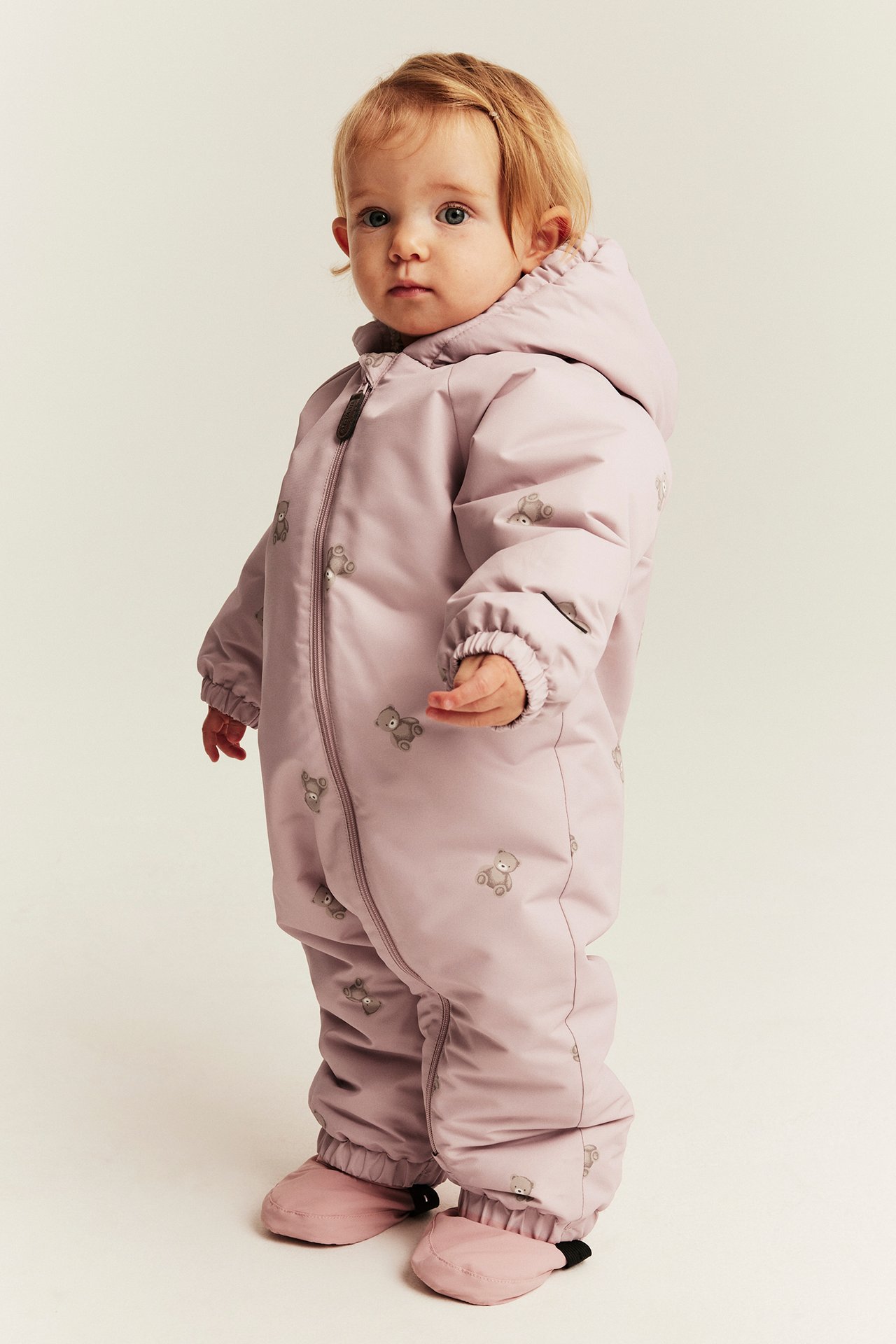 Baby wears a light purple padded snowsuit with hood, front zipper, teddy bear print, and booties.