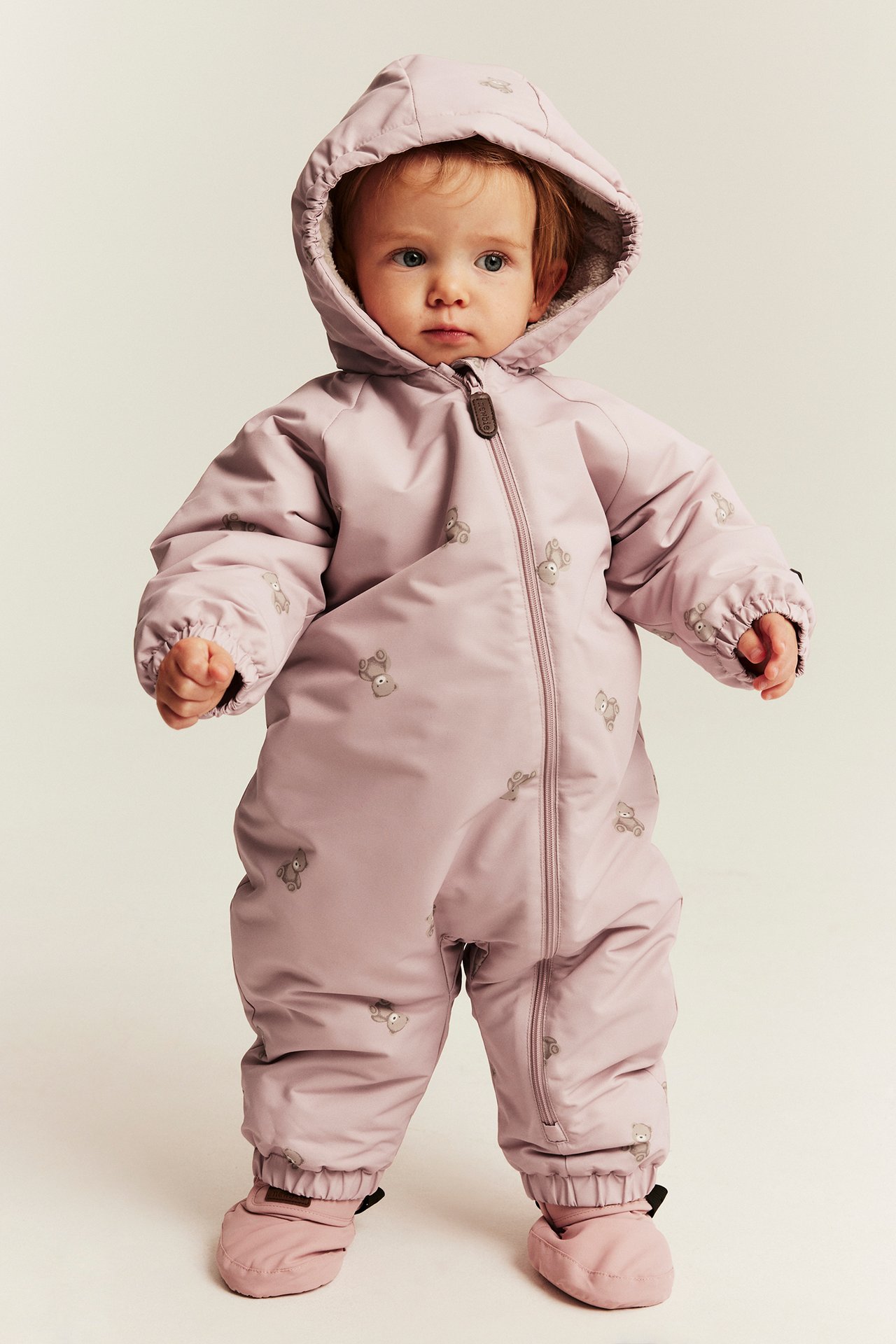Vorderansicht eines Babys in einem hellvioletten Overall mit Tiermuster und passenden weichen Booties.