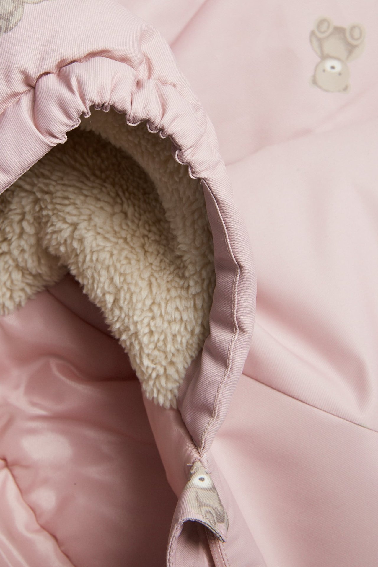 Nahaufnahme eines rosa Babykleidungsstücks mit cremefarbenem Kunstfellfutter und hellgrauer Bärenstickerei.
