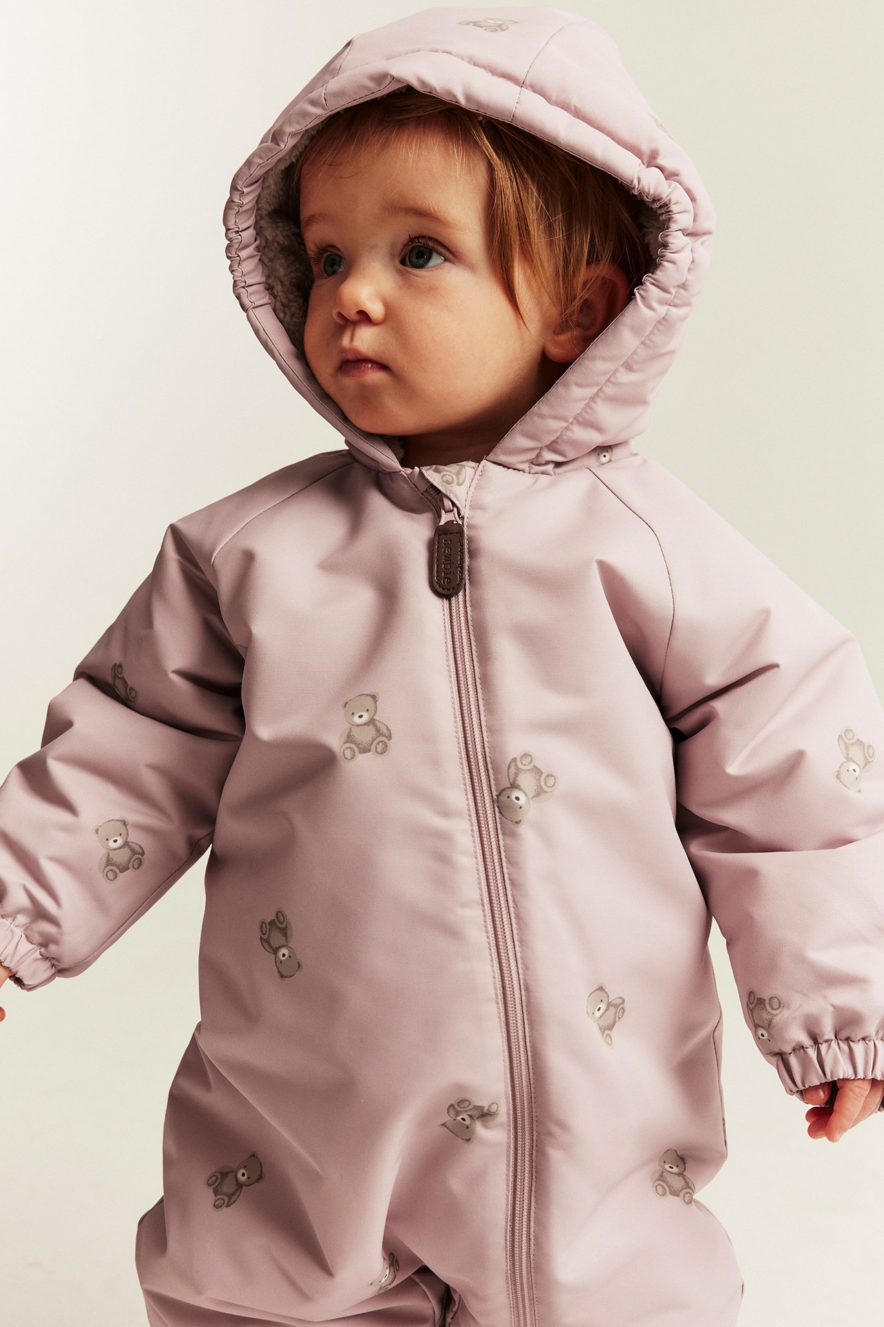 Close-up front view of a baby in a hooded light purple teddy bear print overall with a front zipper.